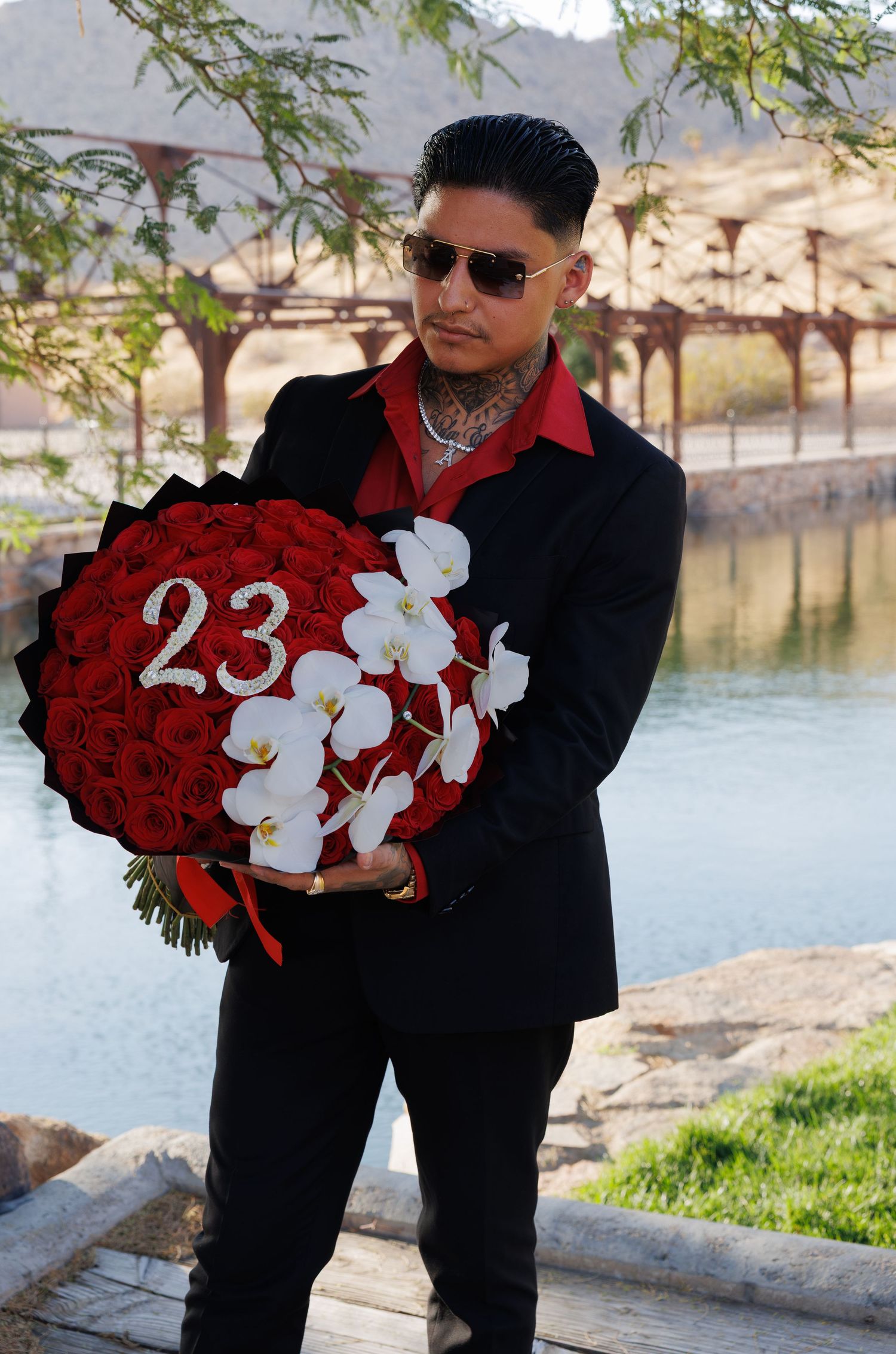 Tattooed man in black suit holding large bouquet of red roses with number 23 outdoors.