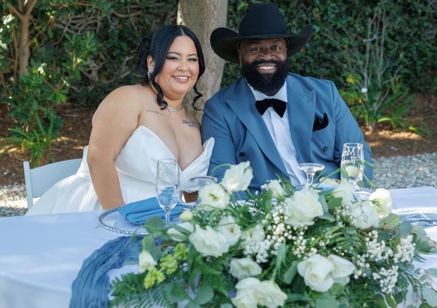 Bride and groom smiling at sweetheart table with white floral centerpiece at outdoor wedding reception.