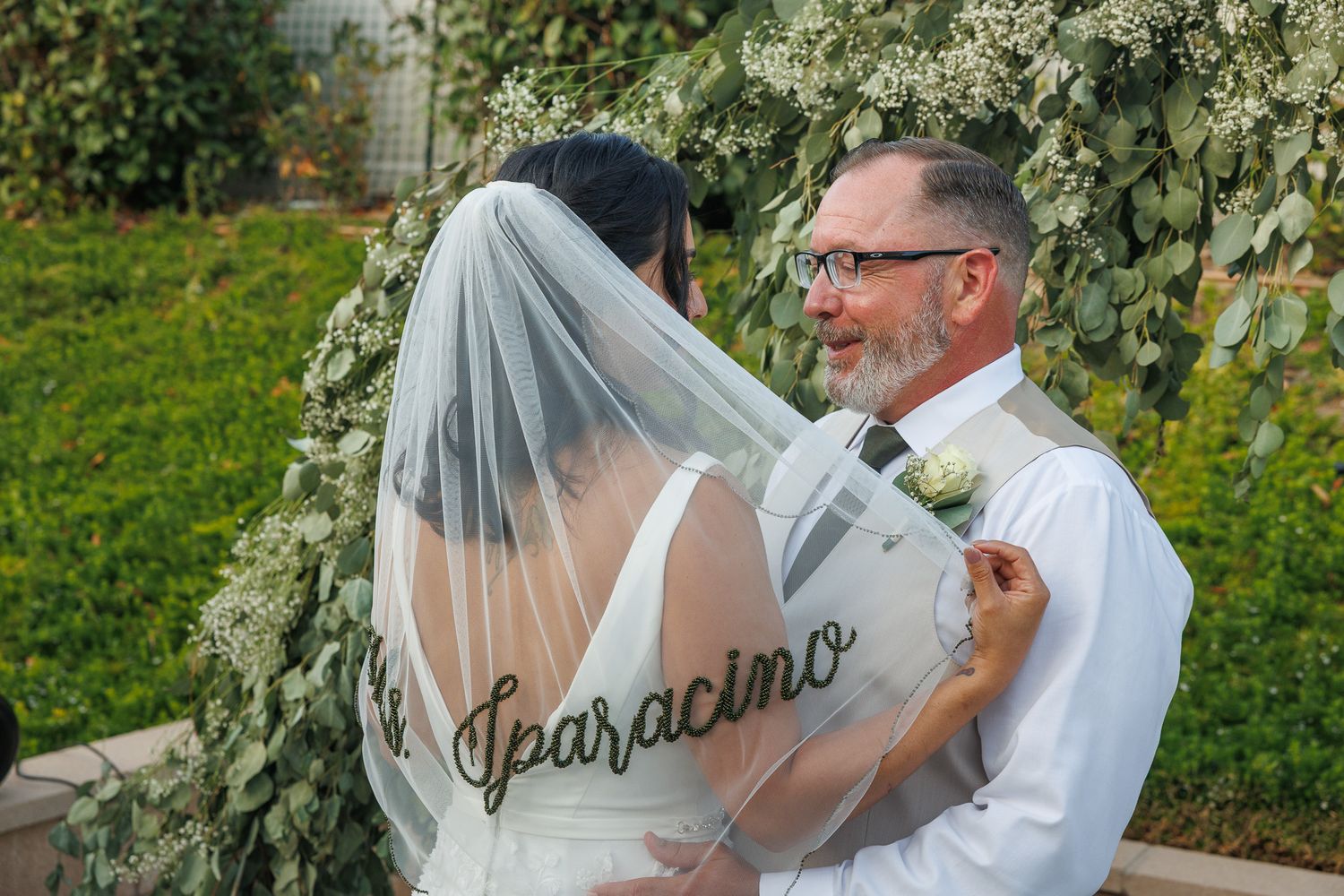 Bride and groom share a tender moment at wedding altar adorned with lush greenery, bride's veil reads Iparacino.