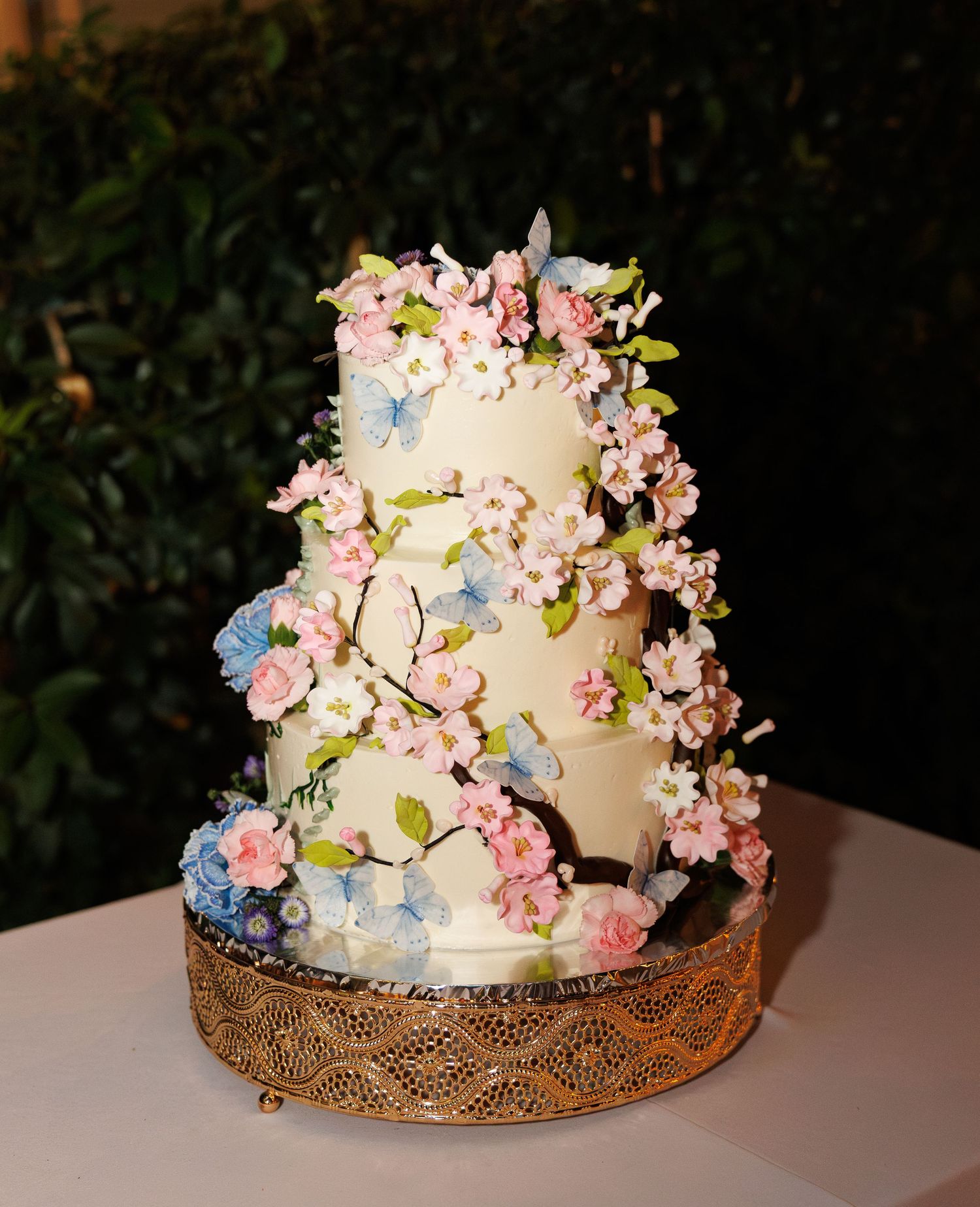 Three-tier white wedding cake adorned with pink cherry blossoms and blue hydrangeas on a gold stand.