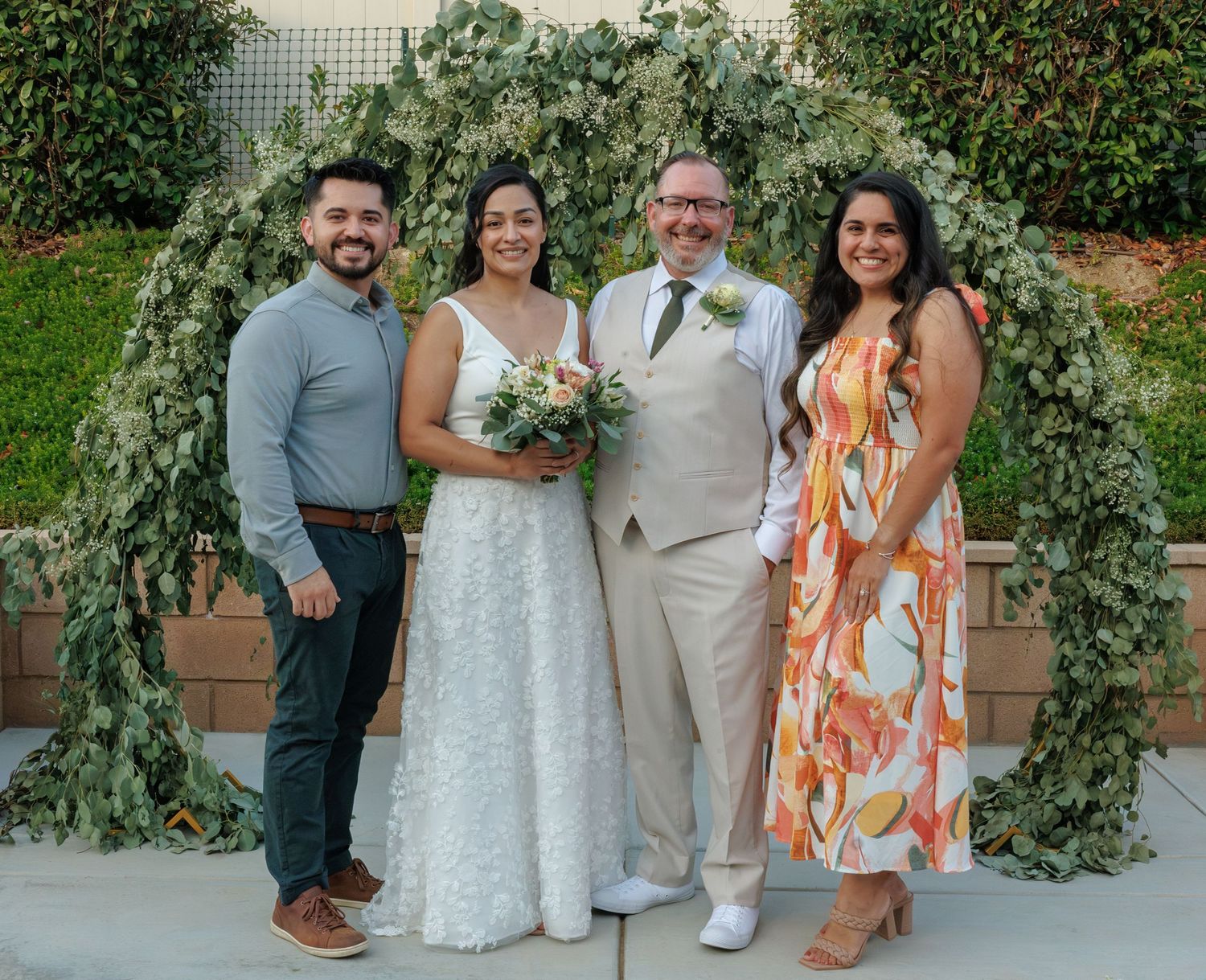 Bride and groom pose with two guests under a beautiful eucalyptus arch at an outdoor wedding.