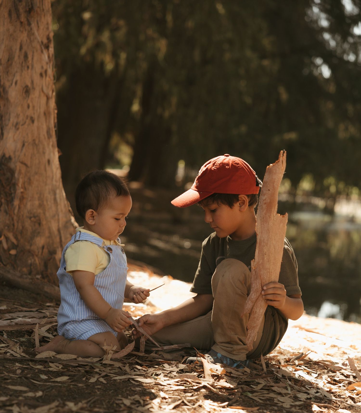 Two young children sit under a tree outdoors, playing with sticks and wood on a sunny day.