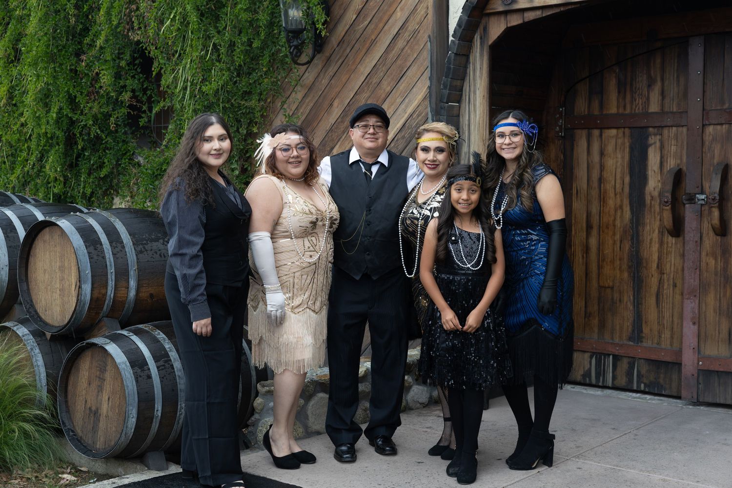 Group of six people dressed in 1920s Great Gatsby themed attire posing together near wine barrels at an event.