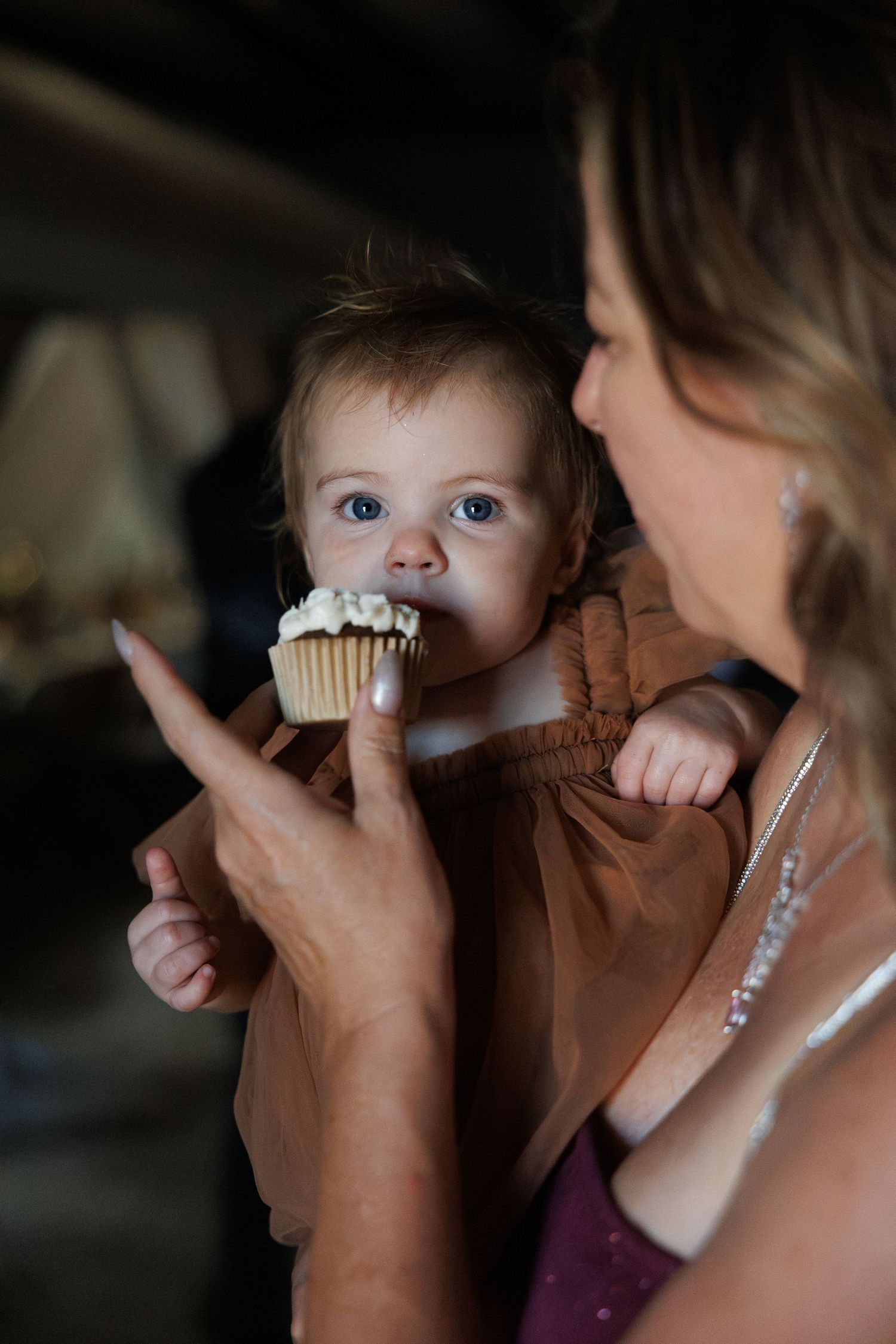 Adorable blue-eyed baby enjoys a cupcake while being held by a woman at a wedding celebration.