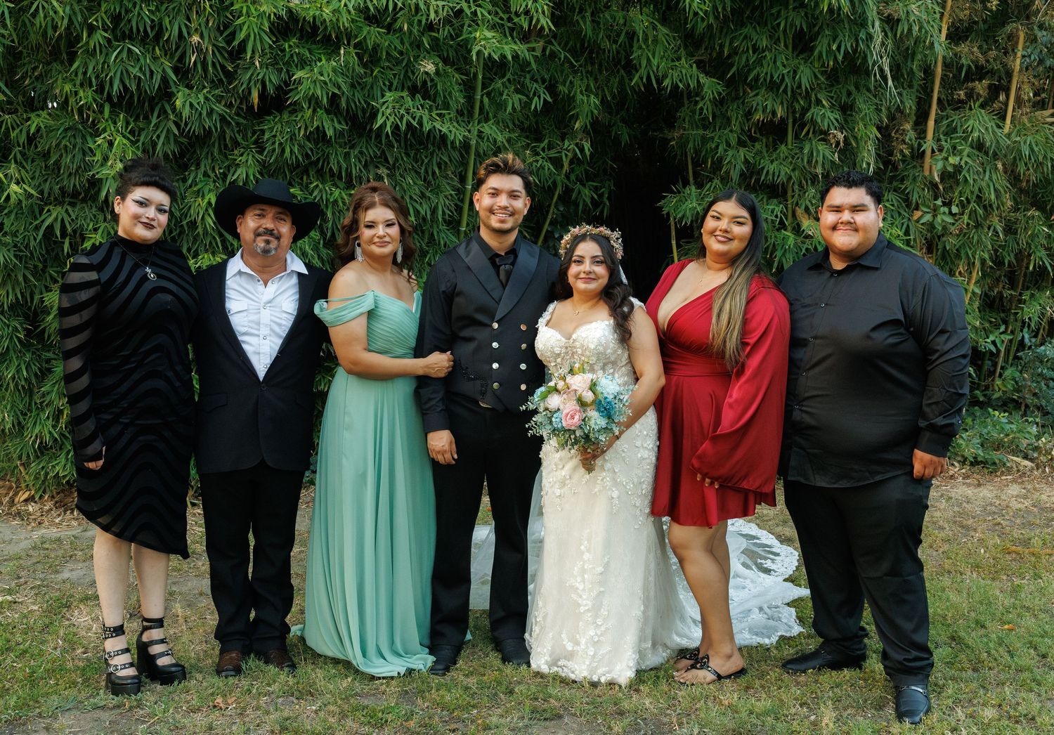 Wedding party poses outdoors with bride in white lace gown and groom in black suit with family.