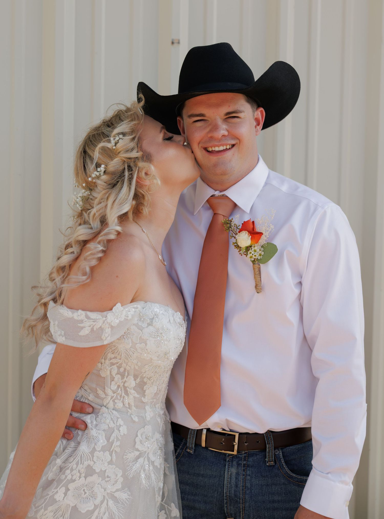 A bride in a lace gown kisses a groom wearing a black cowboy hat, white shirt, and boutonniere at a country wedding.