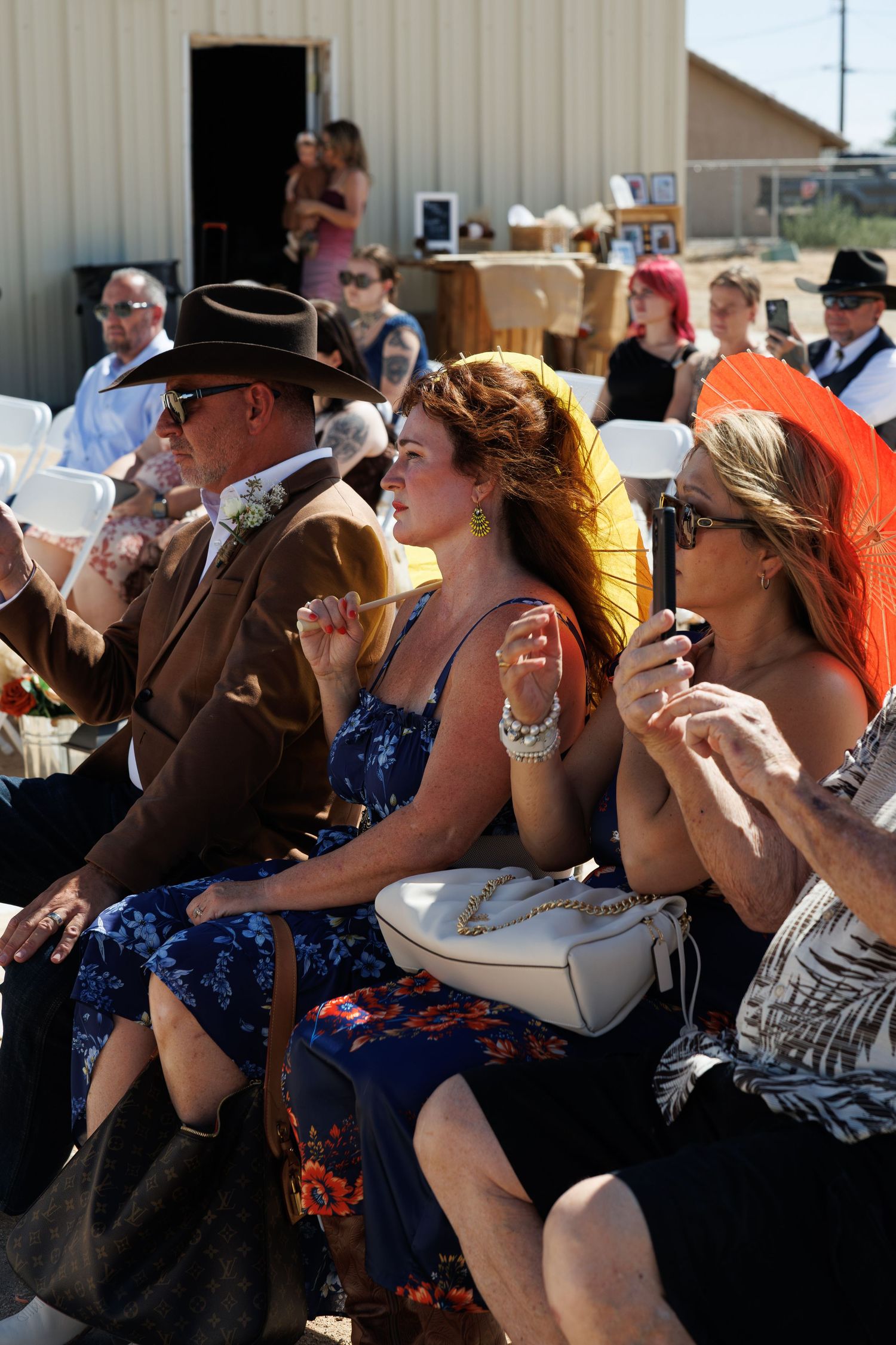 Wedding guests seated outdoors in colorful attire, fanning themselves in the summer heat.