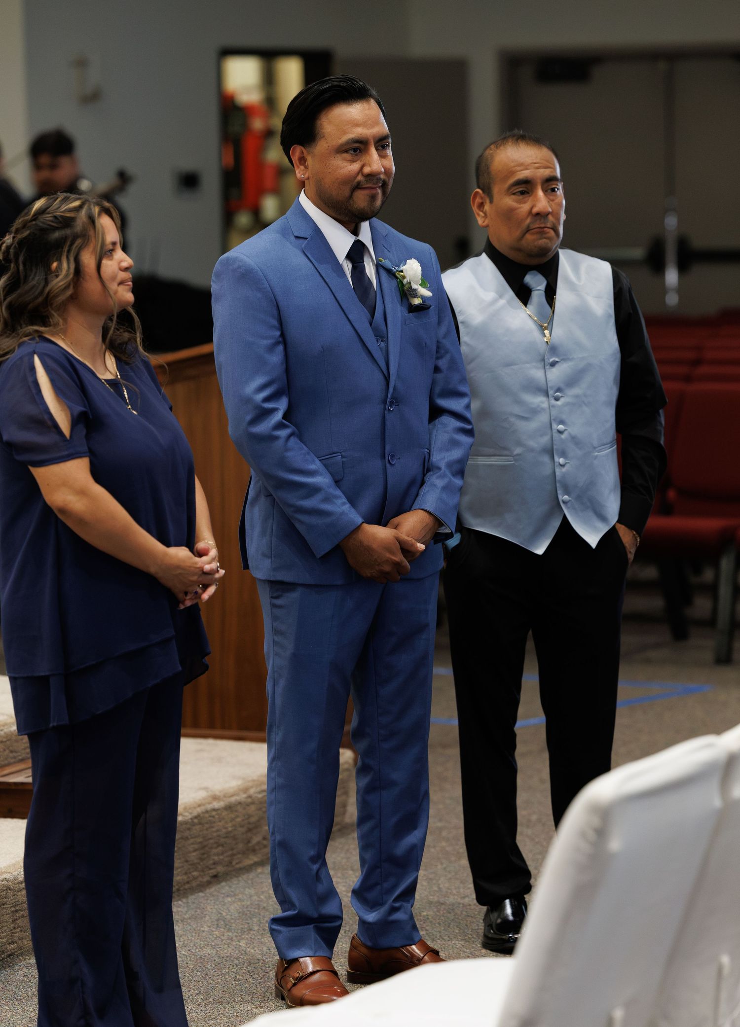 Groom in a blue suit with a boutonniere stands between a woman in a navy outfit and a man in a gray vest during a wedding ceremony.