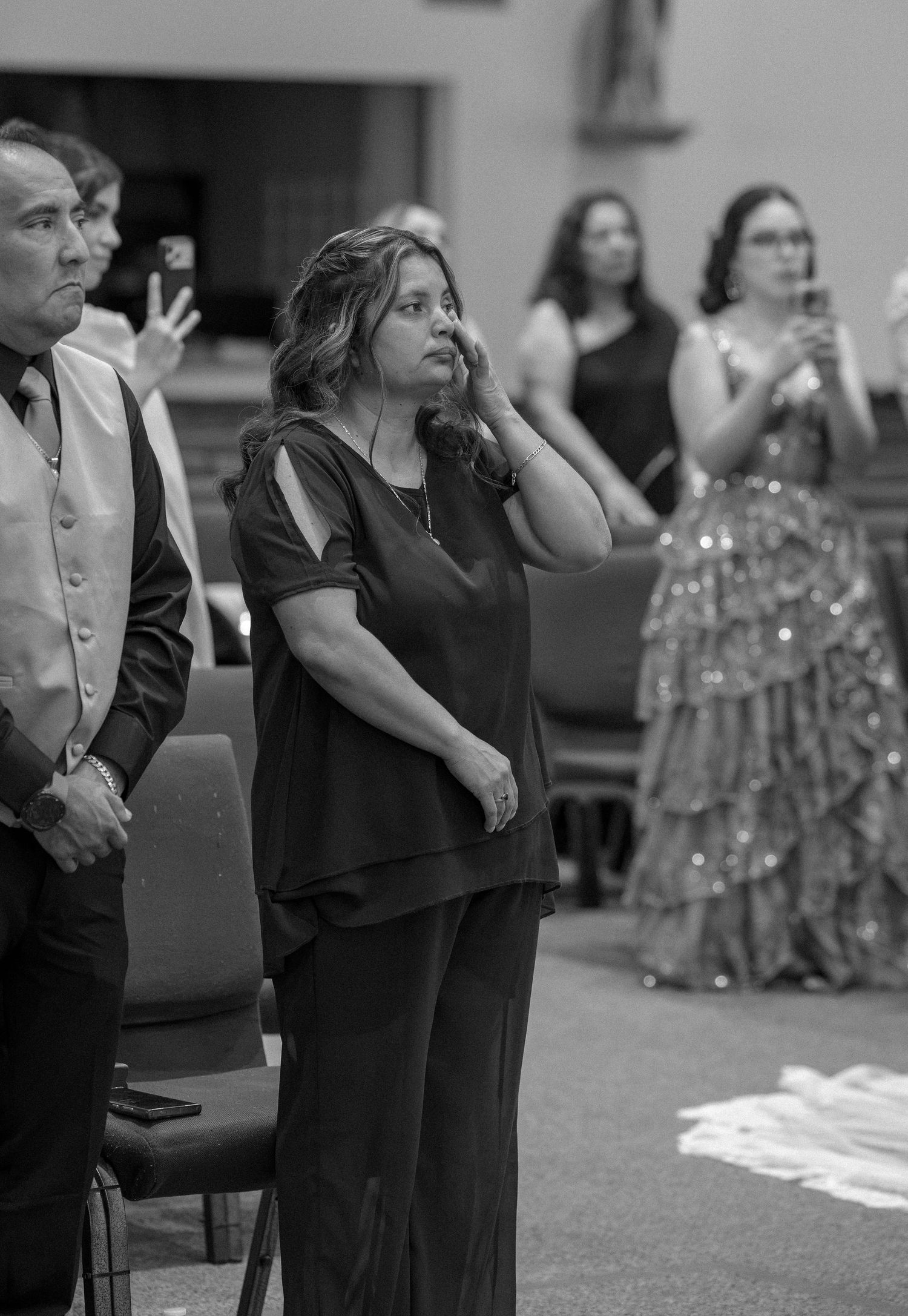 Black and white photo of a woman in dark clothing standing with her hand near her ear during a church ceremony, guests behind her.