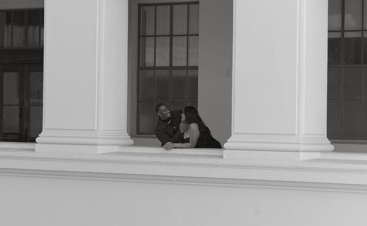 Black and white photo of a couple sitting intimately between white columns on a building ledge.