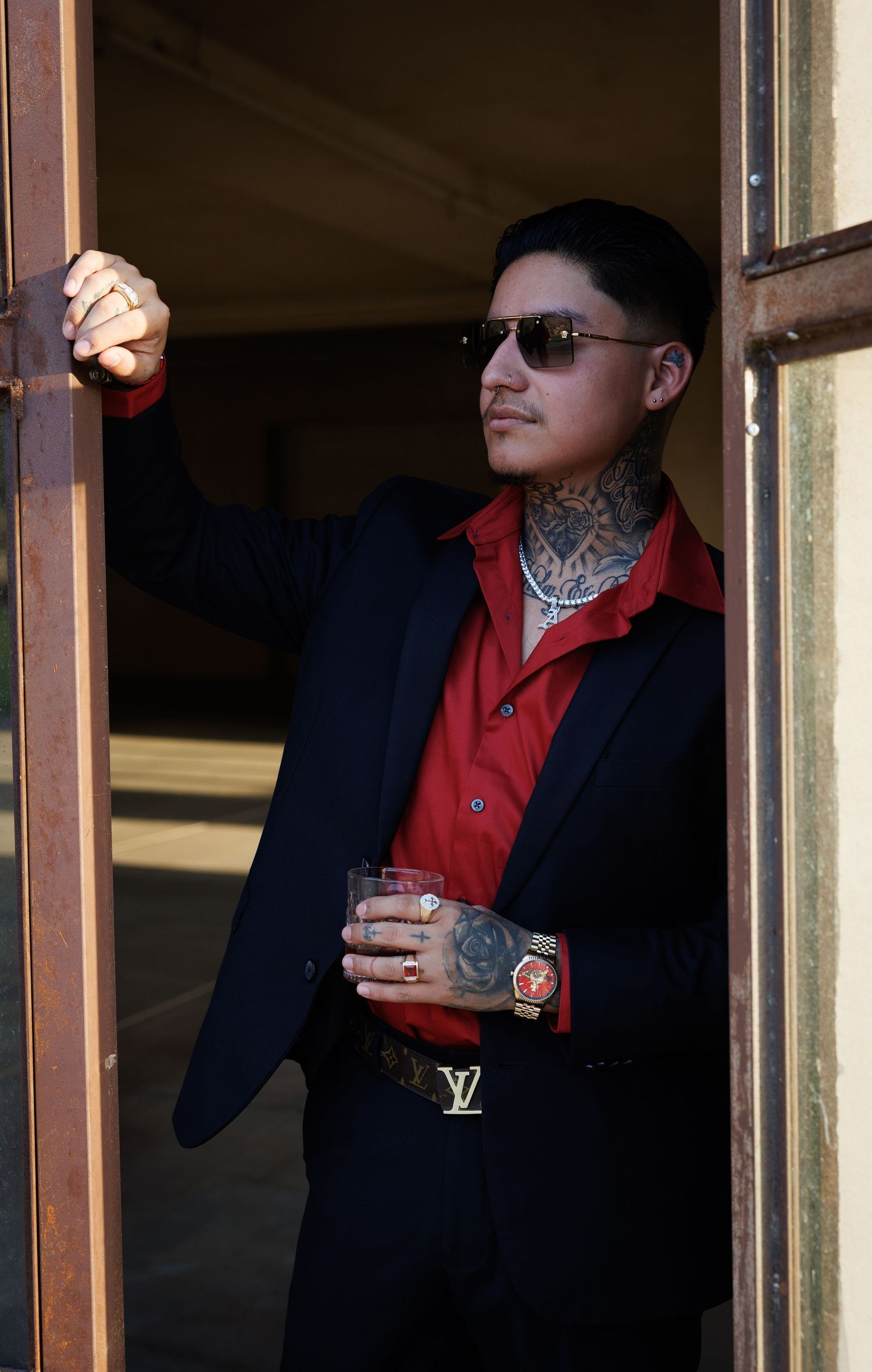 Tattooed man in navy suit and red shirt holding a glass, wearing Louis Vuitton belt and sunglasses, leaning in doorway.