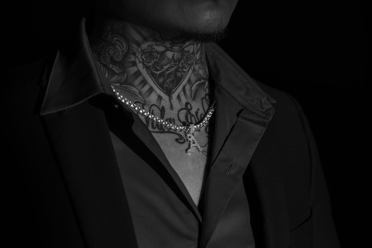 Black and white photo of a person with neck tattoos wearing a suit and silver chain necklace.