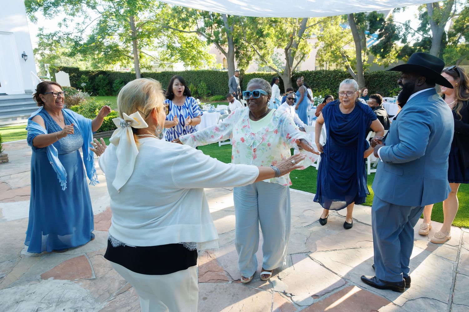 Wedding guests dance together outdoors under white canopy during sunny daytime reception celebration.