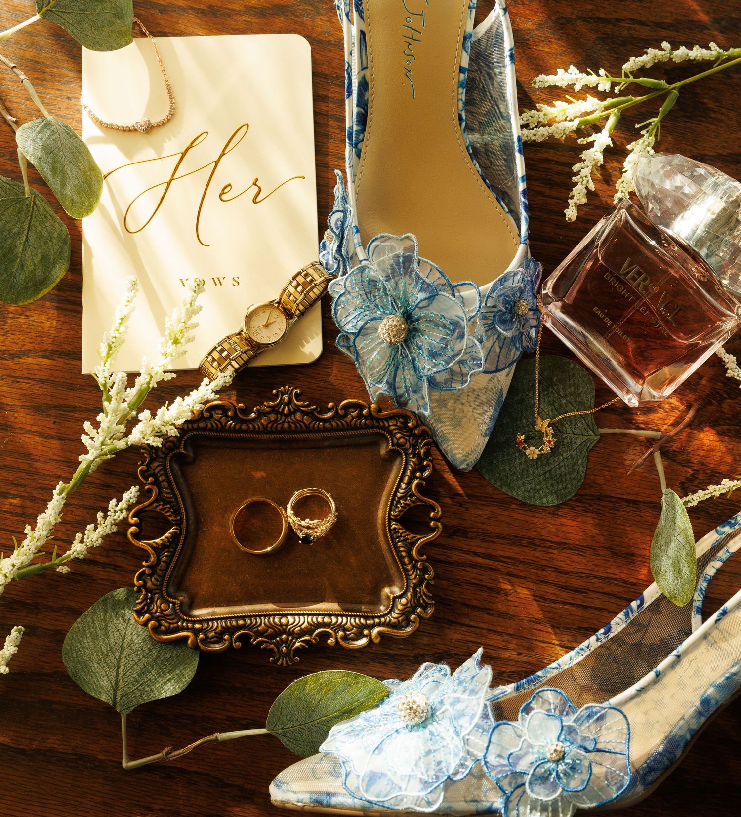 Elegant bridal flat lay with blue floral heels, gold rings, vow book, watch, eucalyptus, and perfume on wood.