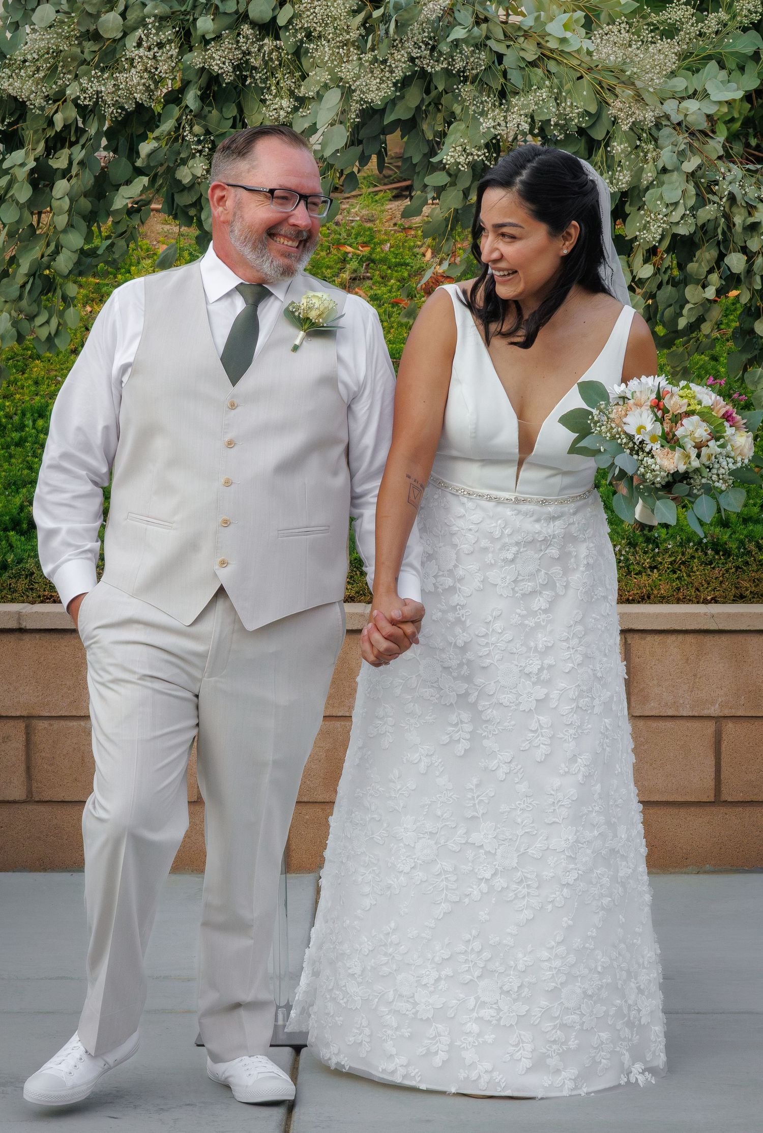 A smiling bride in a white lace gown holds a bouquet beside her groom in a tan suit with boutonniere outdoors.