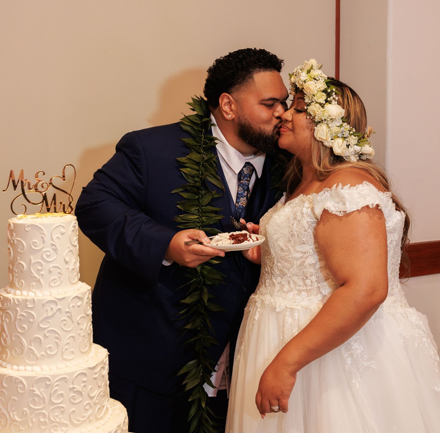 Newlyweds share a kiss while cutting their white tiered wedding cake, groom in blue suit with lei, bride in white gown.