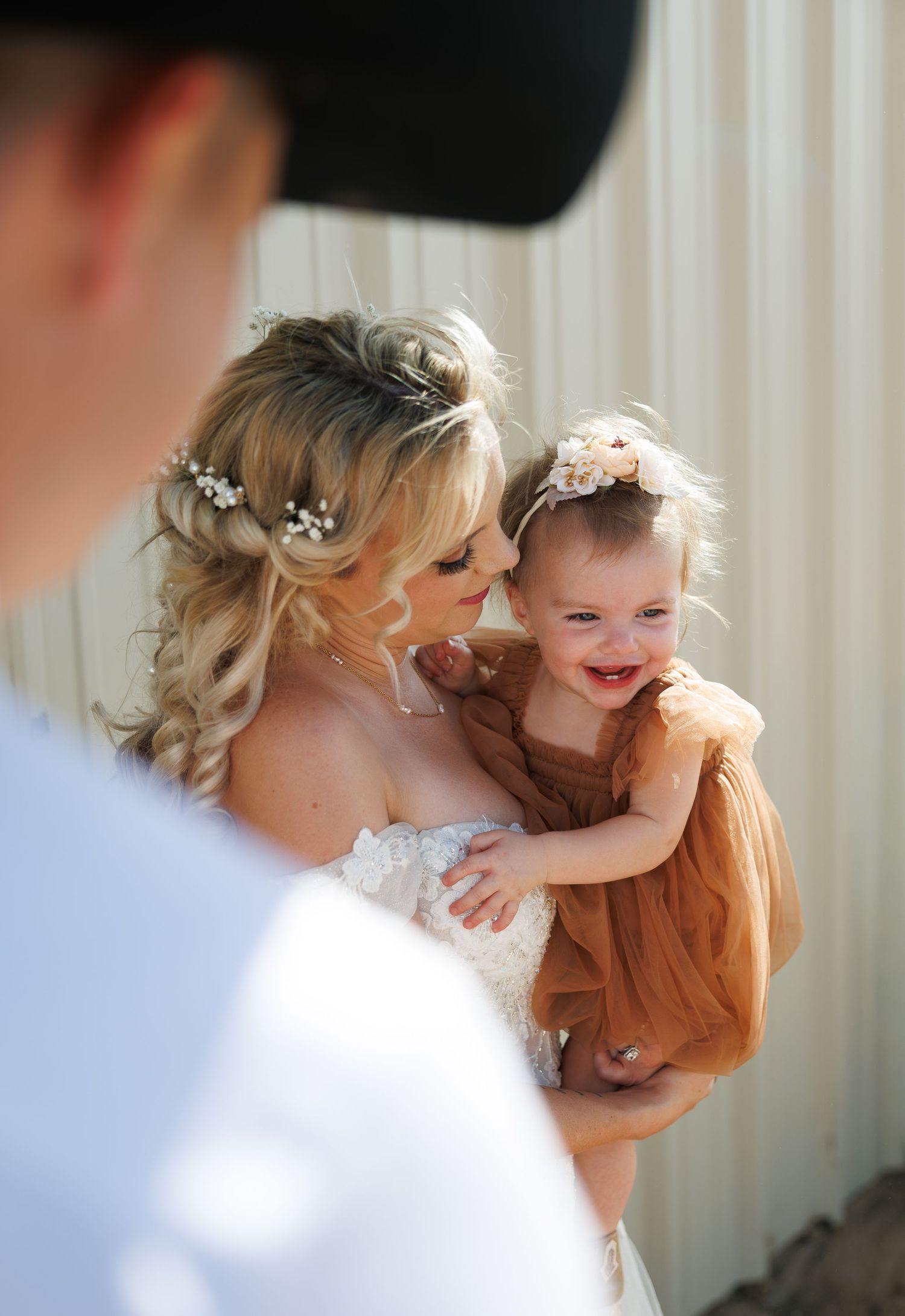 Bride in lace gown with floral hair accessories holds smiling baby girl in rust tulle dress at outdoor wedding.