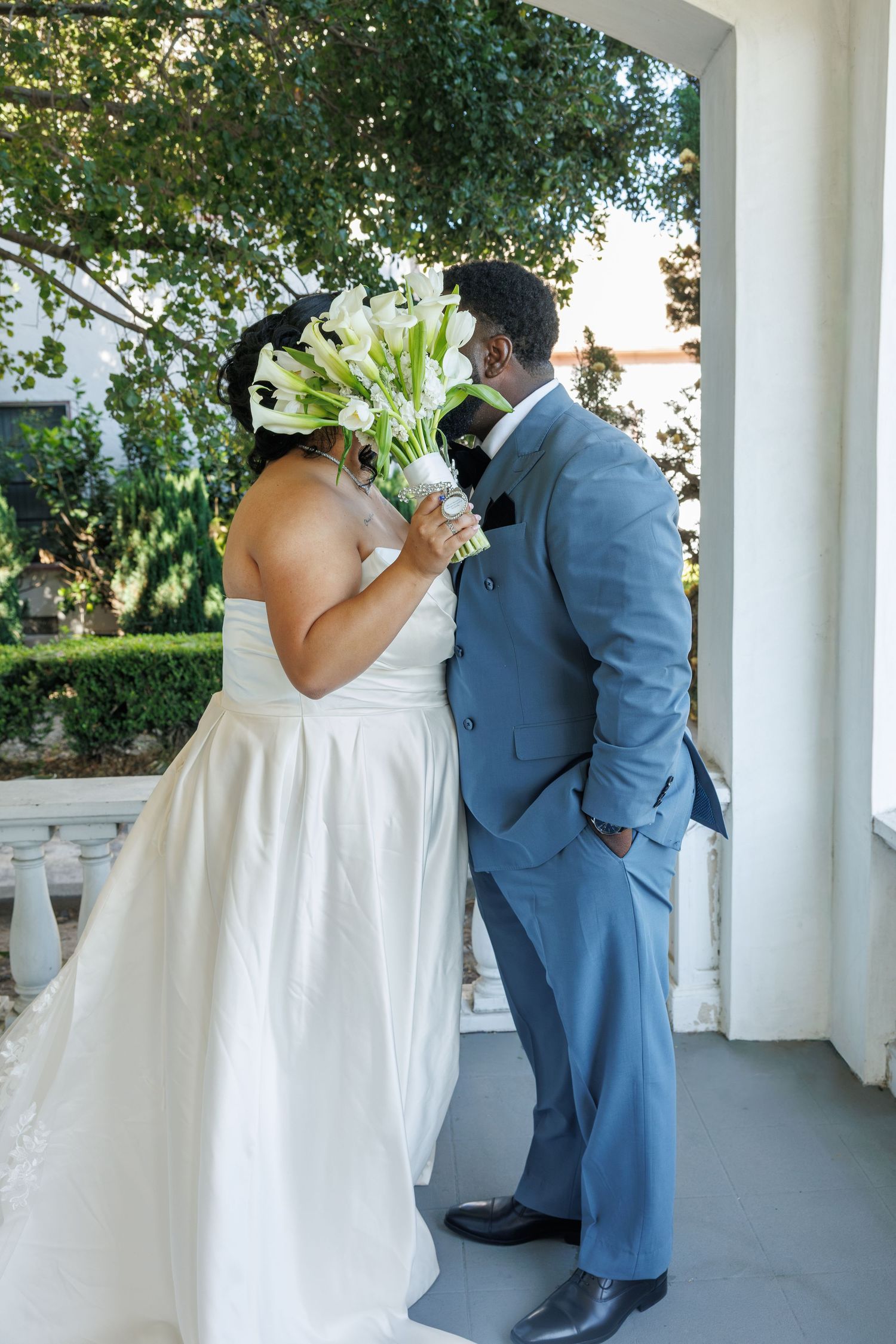 Bride in white gown holds calla lily bouquet while kissing groom in blue suit on elegant venue porch.
