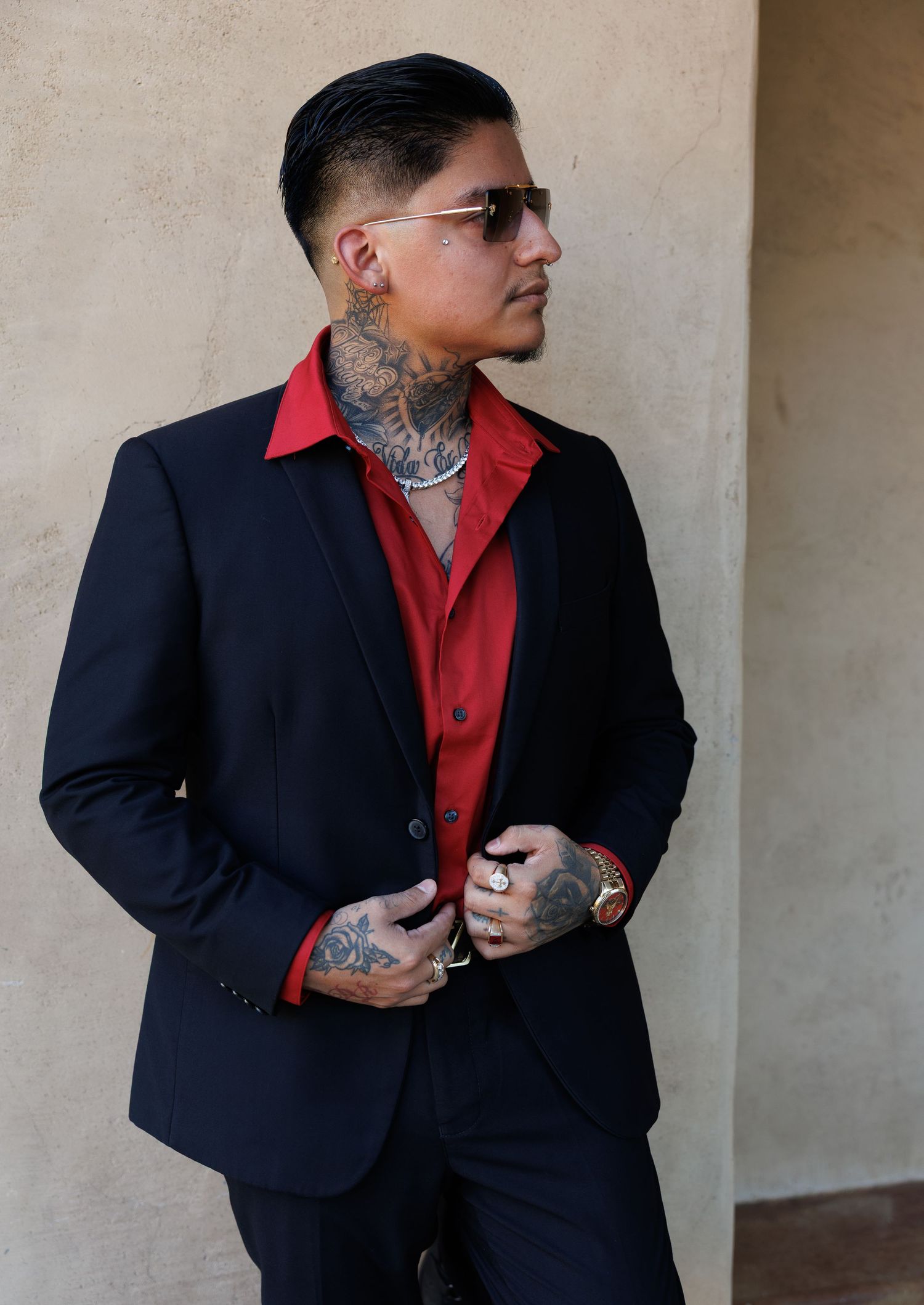 Tattooed man in black suit and red shirt with sunglasses posing outdoors in stylish portrait.