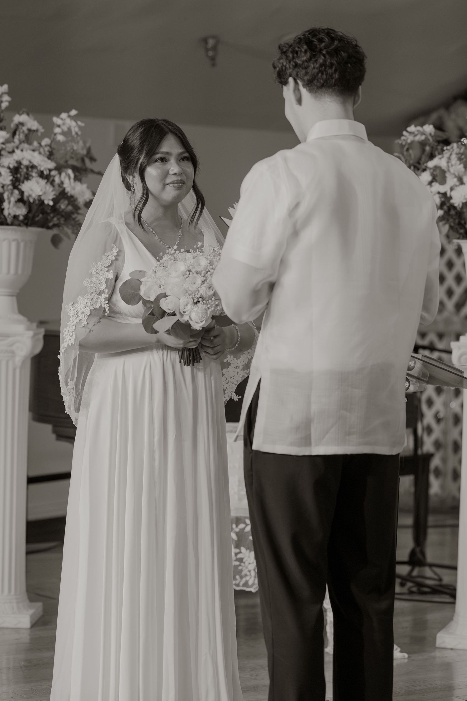 Black and white photo of a bride and groom exchanging vows at a wedding ceremony with floral arrangements.