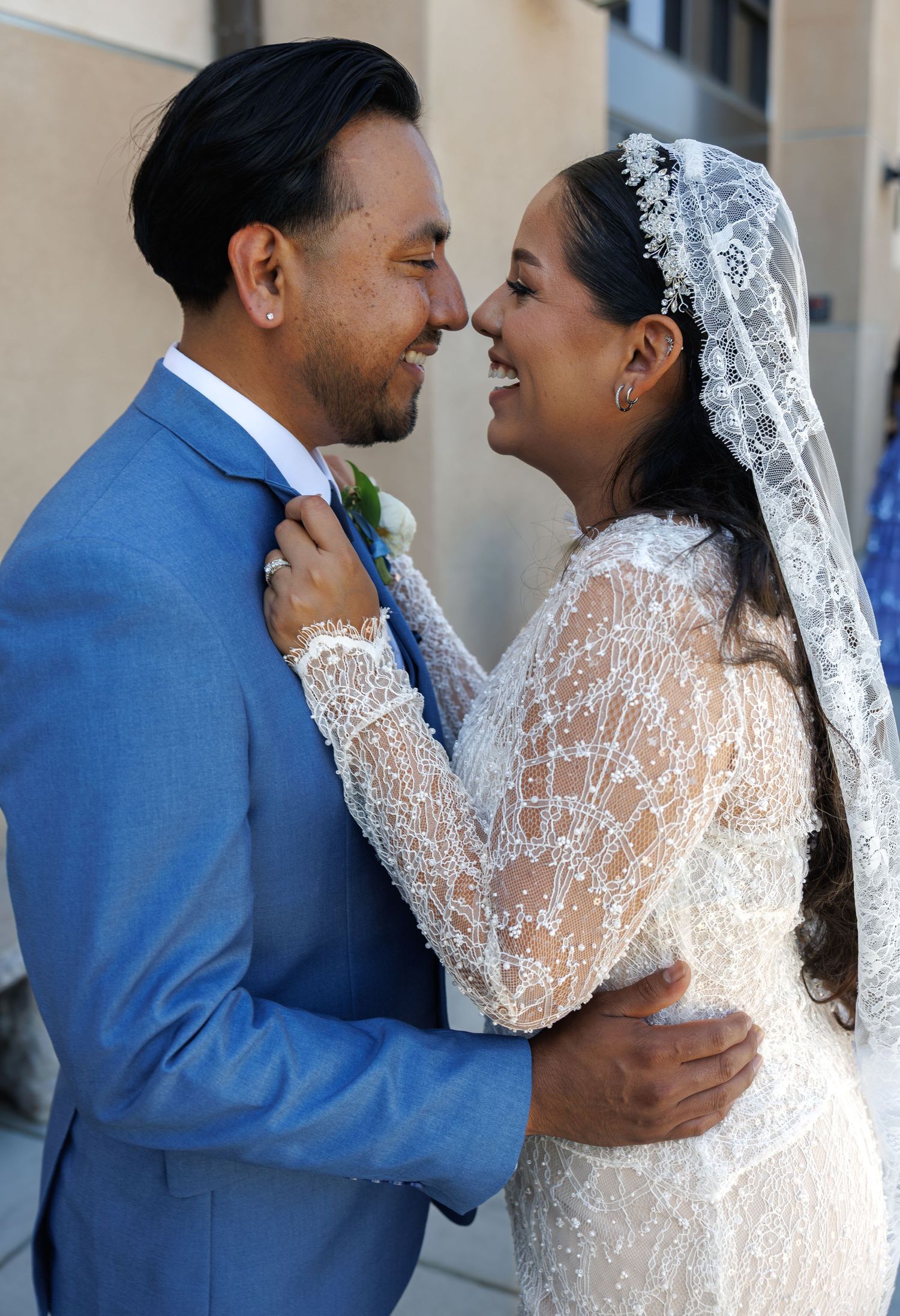 Smiling bride in a beaded lace wedding gown and veil shares a tender moment with her groom in a blue suit outside the ceremony.