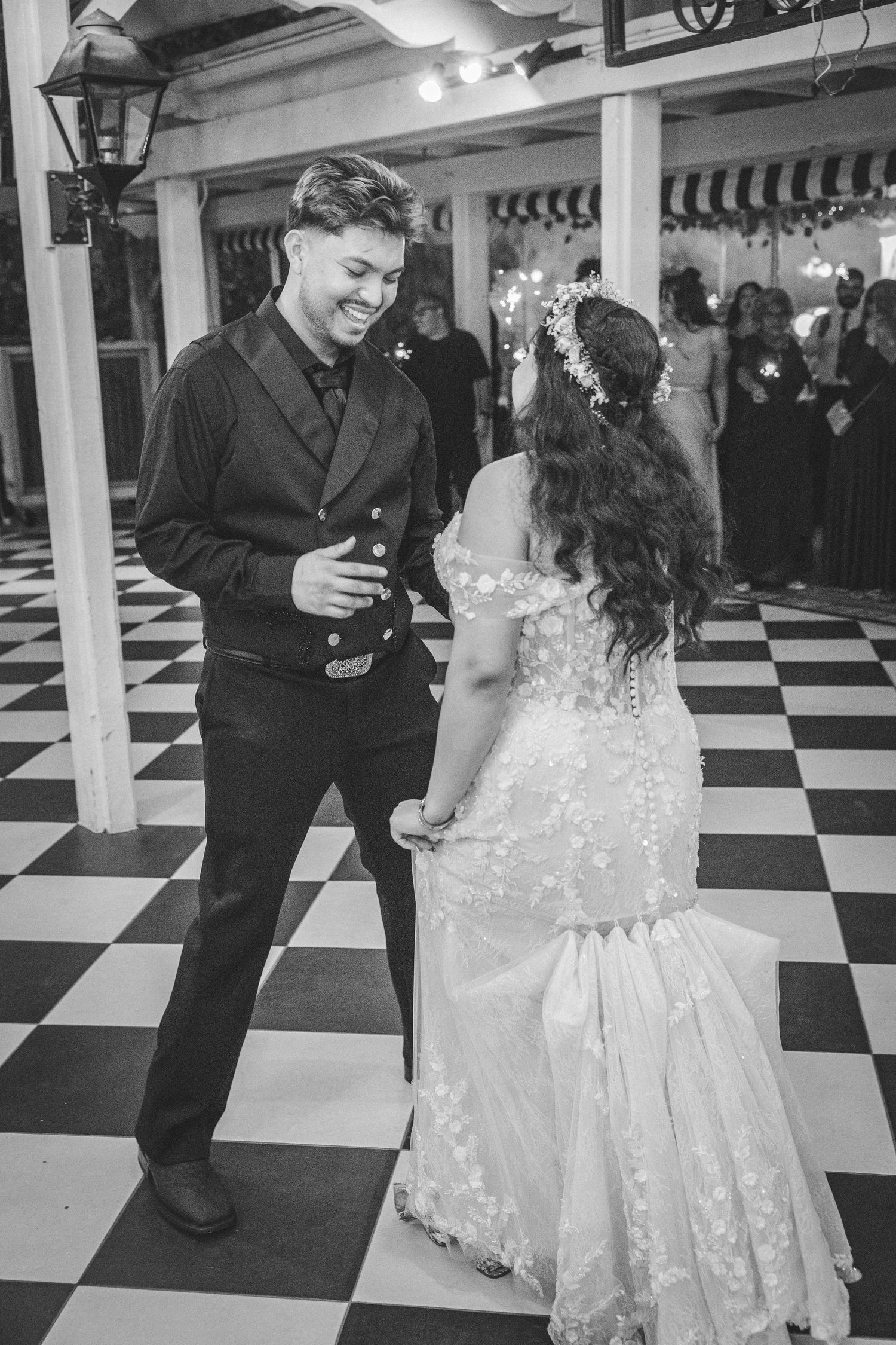 Black and white photo of a smiling groom dancing with bride in lace dress on checkered floor at wedding reception.