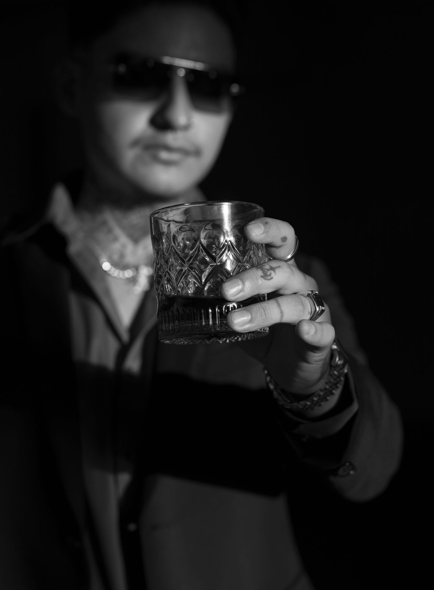 Tattooed man in sunglasses holding a crystal whiskey glass in a moody black and white portrait.