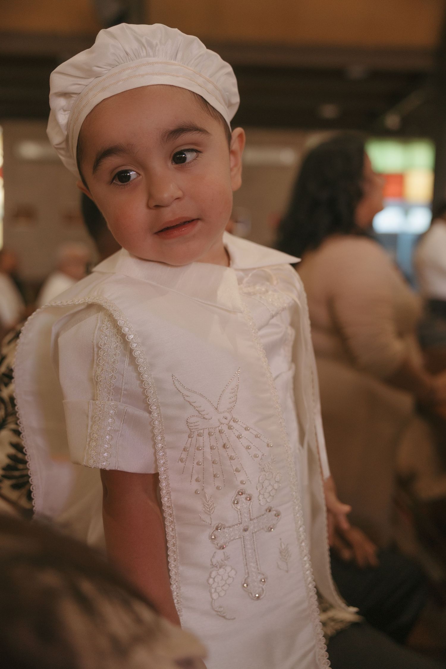 Young child in white baptism outfit with embroidered cross design and matching hat at a religious ceremony.