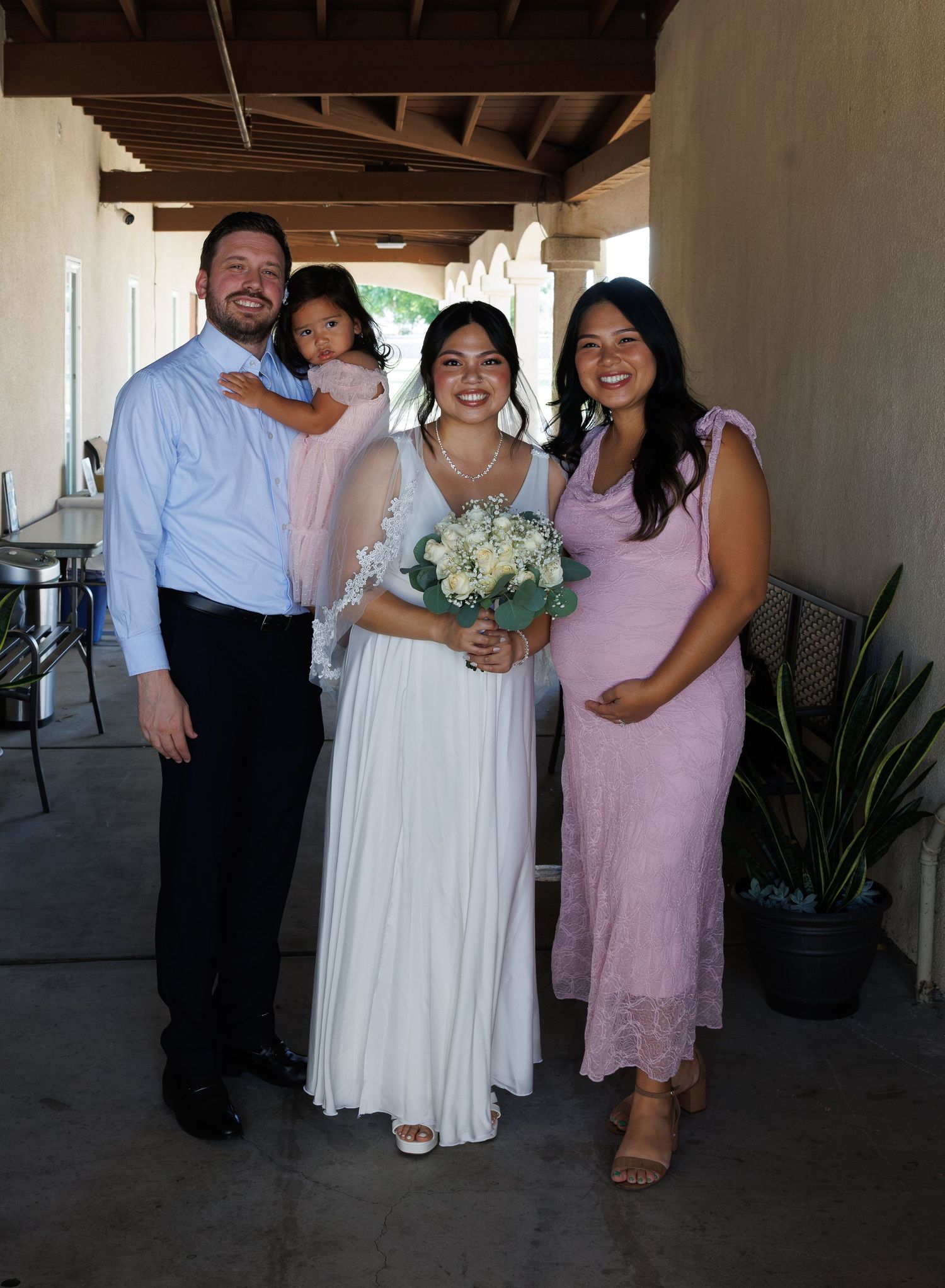 A bride in a white gown holds a bouquet, posing with family including a pregnant woman in pink at an outdoor wedding.