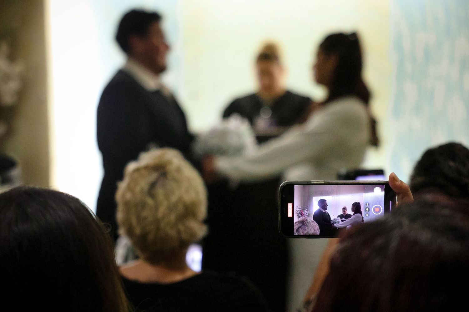Guest records wedding ceremony on smartphone as couple stands with officiant during indoor vows.