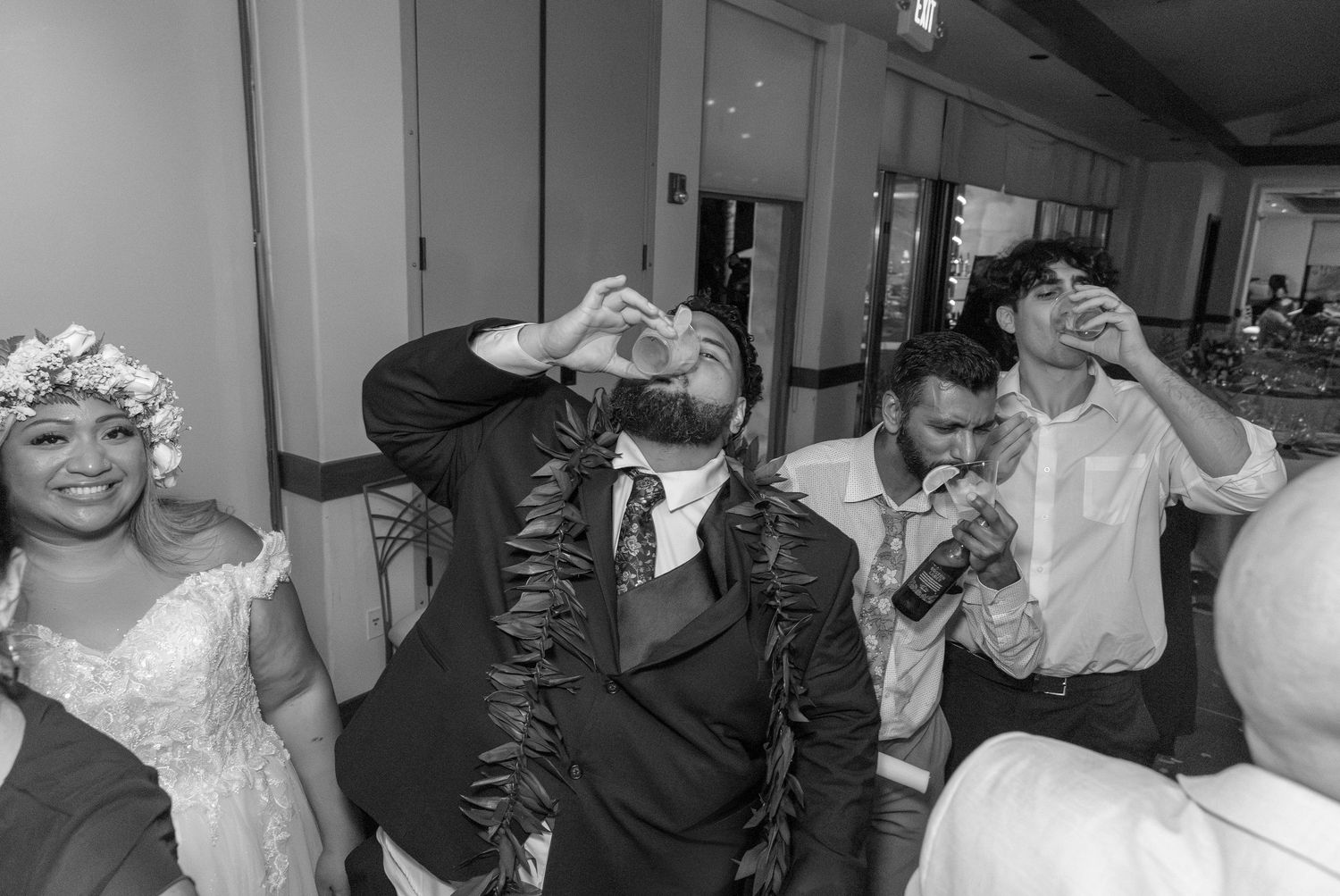 Groom drinks a shot at wedding reception while guests celebrate around him in black and white photo.