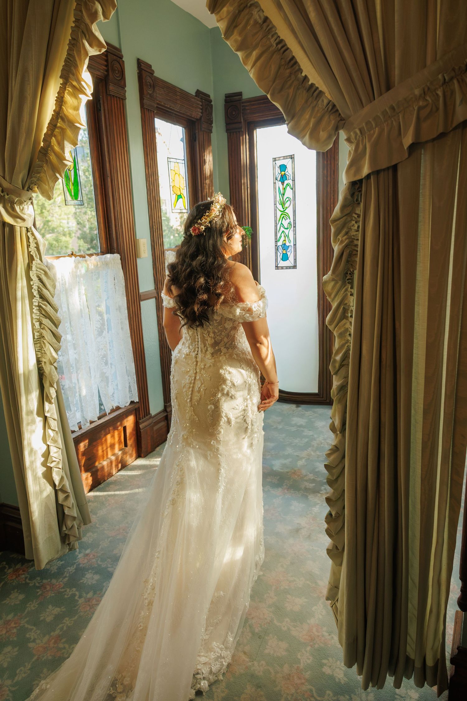 Bride in lace off-shoulder wedding gown stands in elegant vintage hallway with stained glass windows and gold drapes.