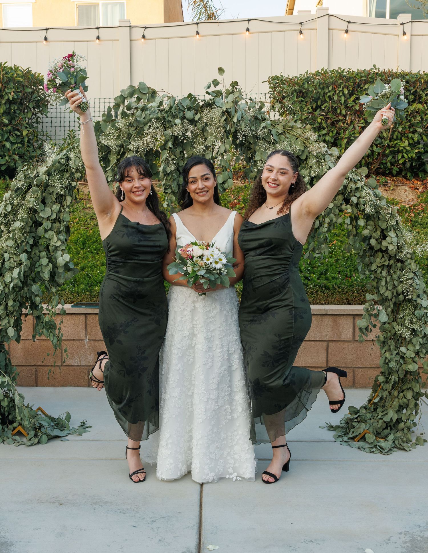 Bride in white lace gown poses with two bridesmaids in olive green dresses under a eucalyptus arch.
