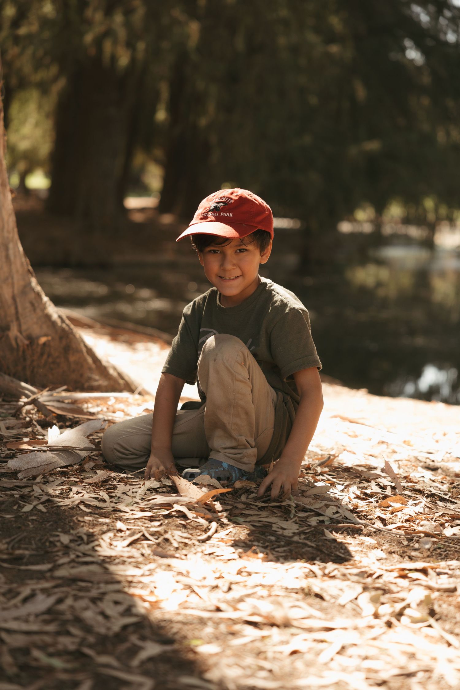 A smiling boy in a red cap sits on fallen leaves in a sunlit park, playing outdoors near trees.