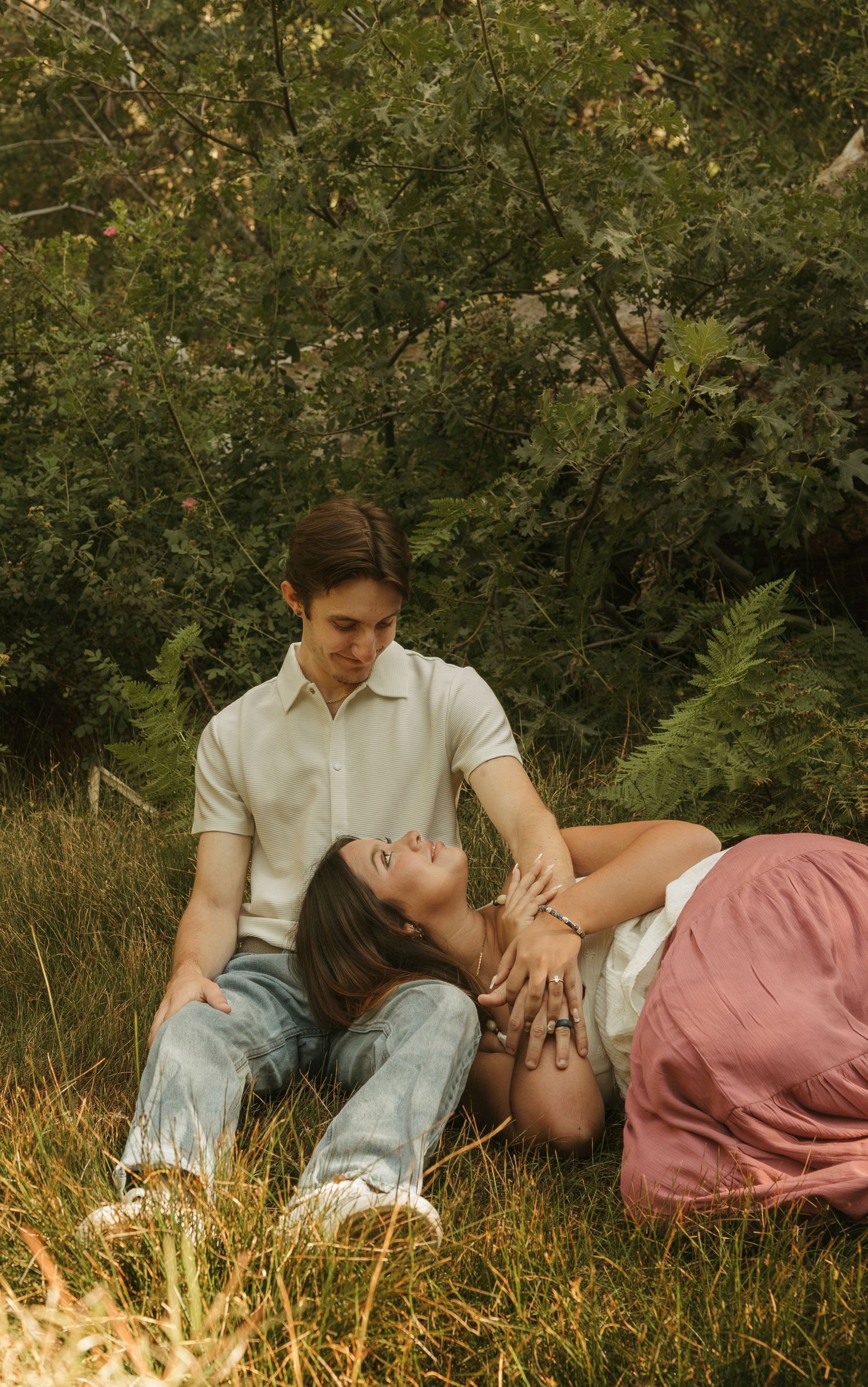 A couple relaxes in a grassy meadow, woman lying in mans lap, surrounded by lush green trees.