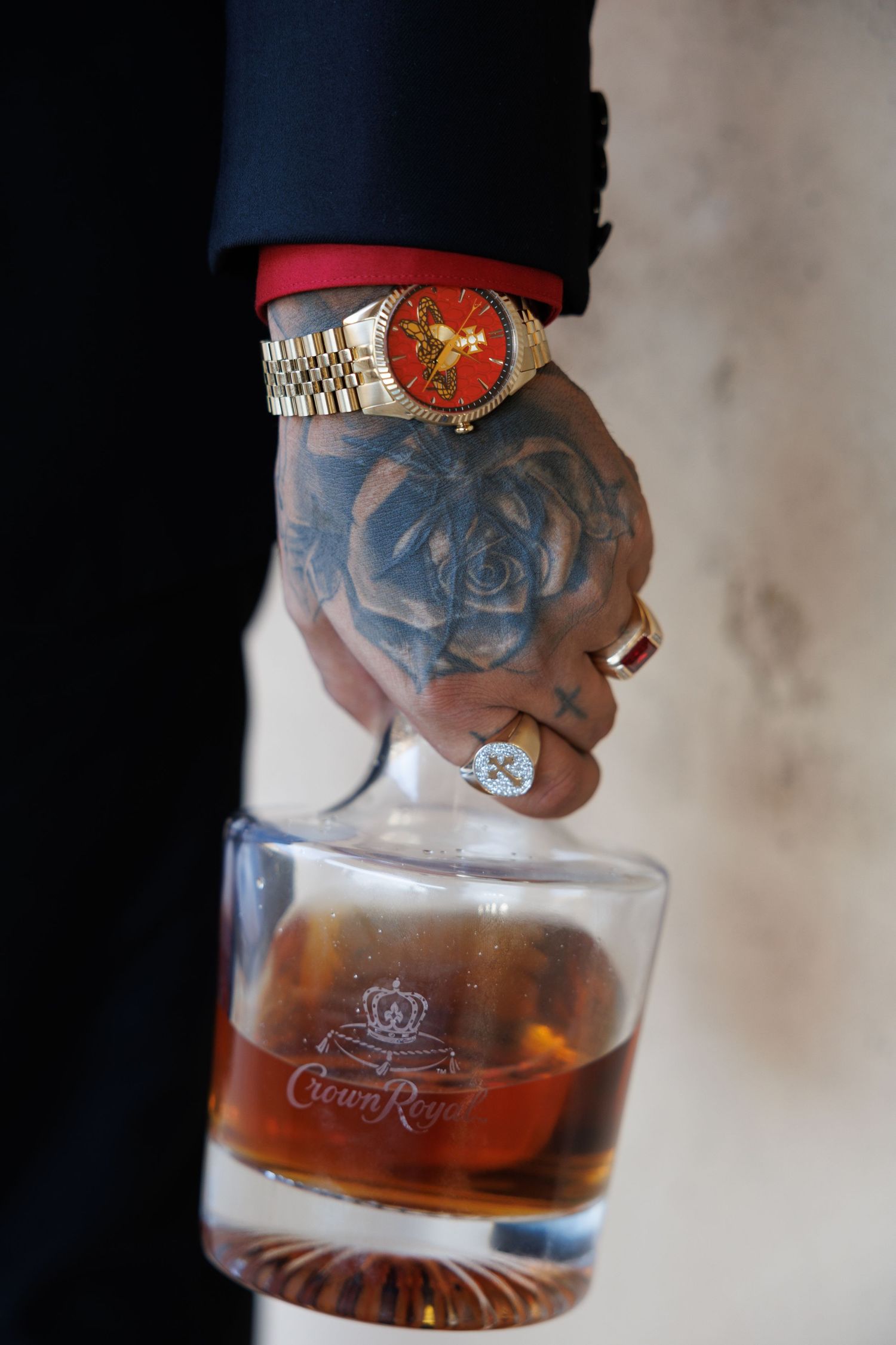 Tattooed hand holding a Crown Royal whiskey glass, wearing a gold watch with red dial and diamond rings.