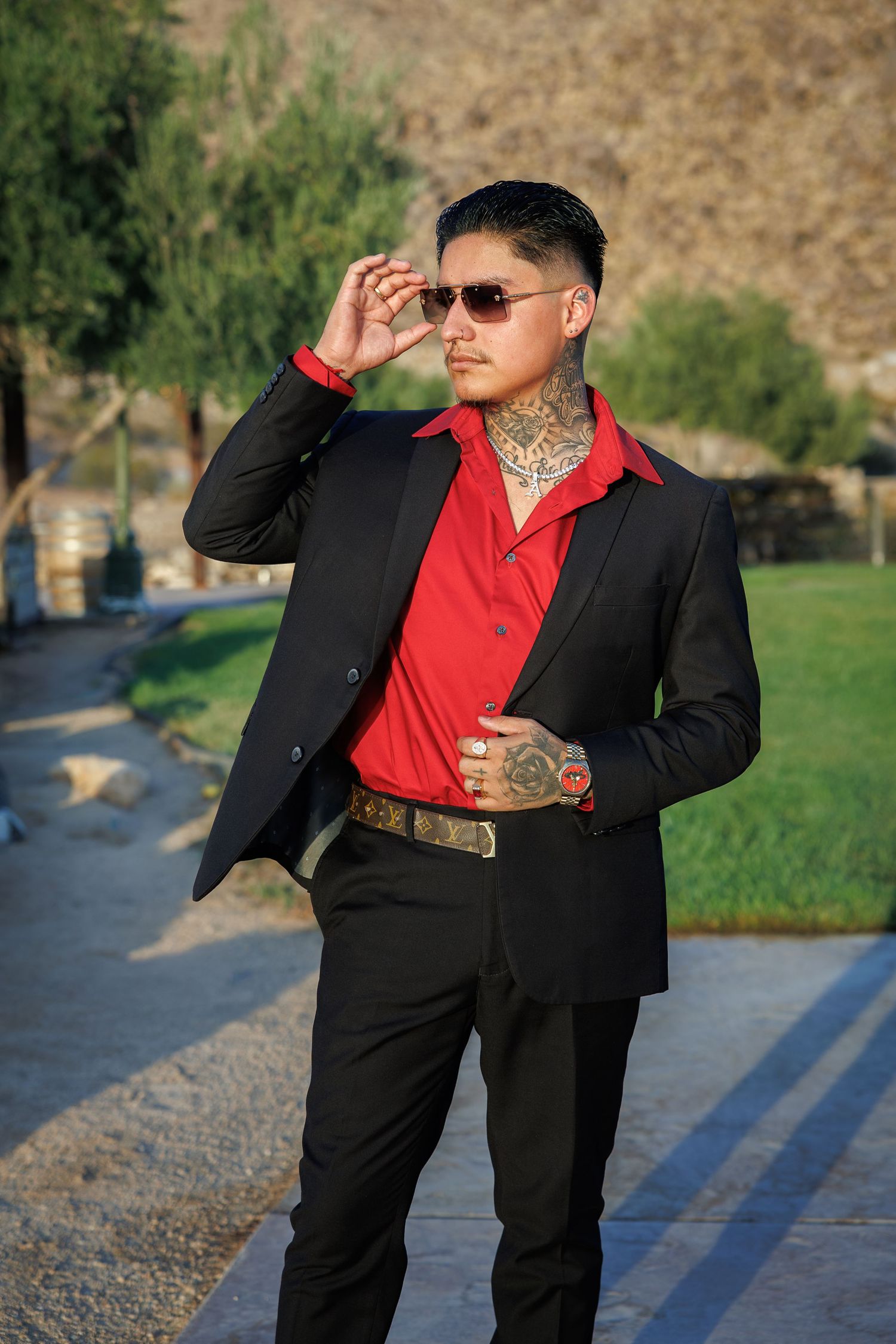 Tattooed man in black suit and red shirt adjusts sunglasses outdoors near green trees and rocky path.