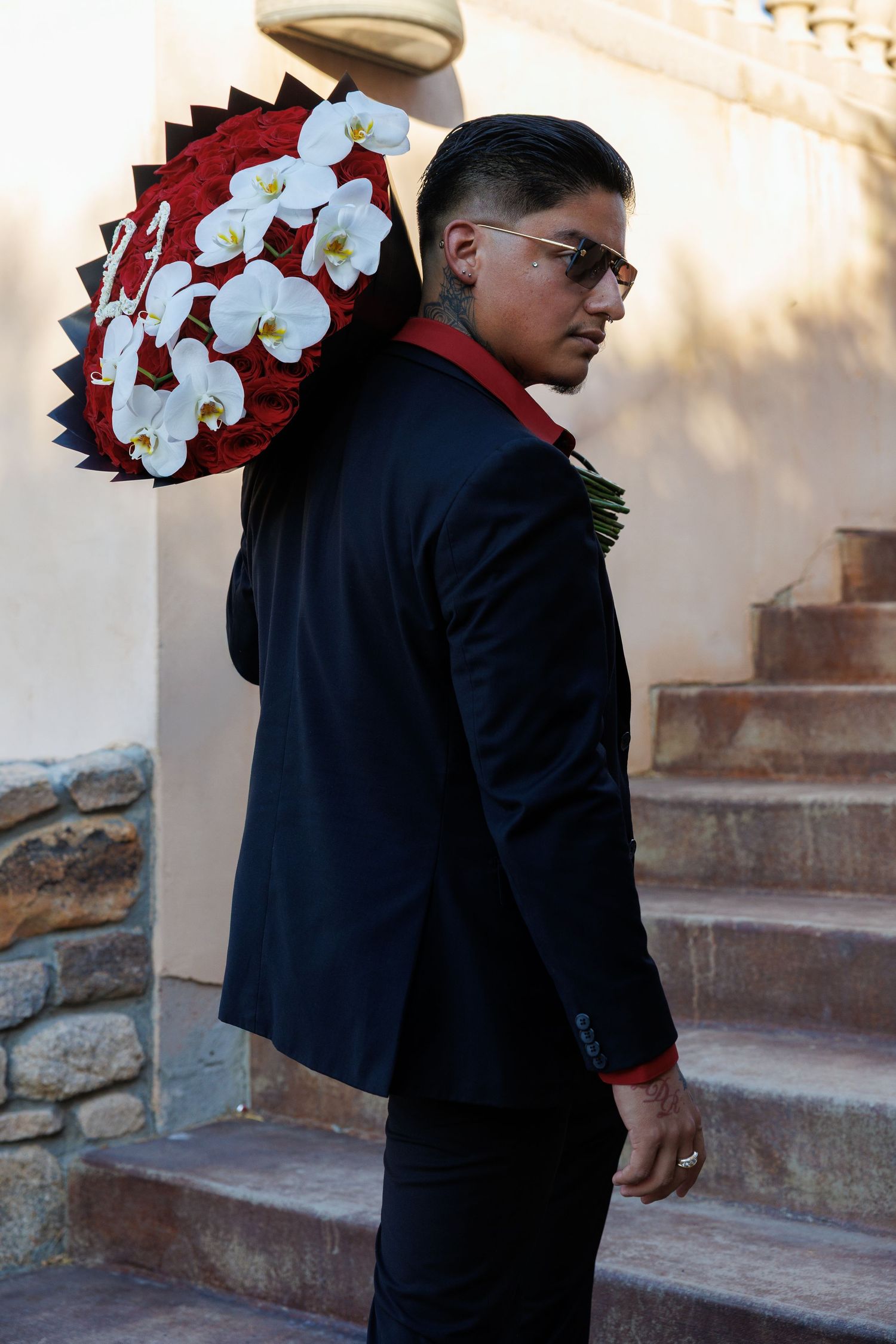Man in black suit holding large red and white orchid bouquet on his shoulder, standing near stone steps.