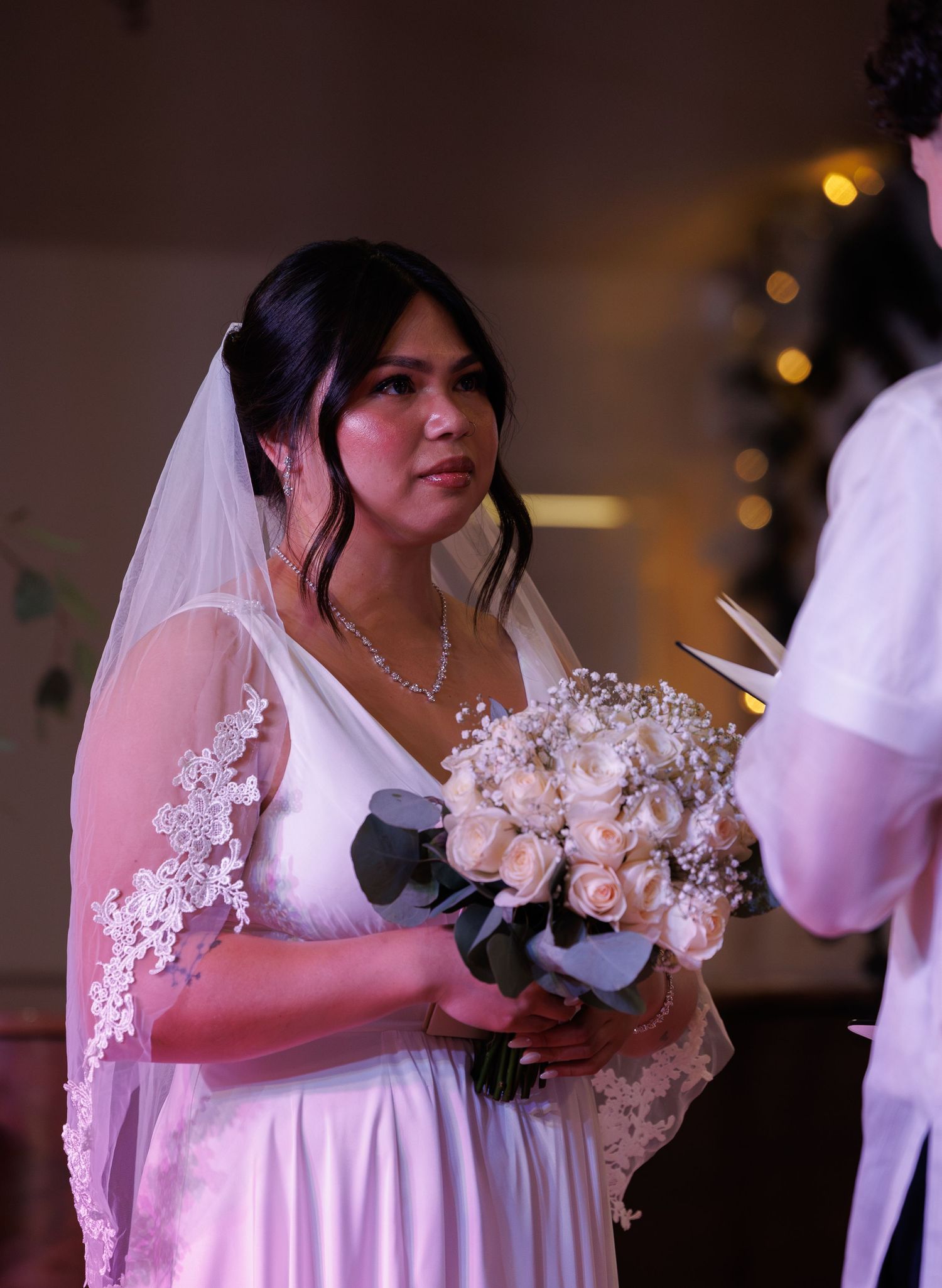 Bride wearing lace veil and white gown holds white rose bouquet during emotional church wedding ceremony.