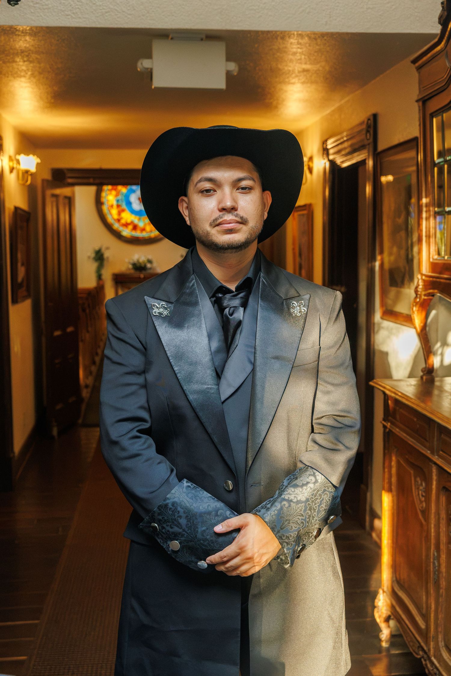 Young man in black cowboy hat and elegant two-tone suit poses in warmly lit hallway with vintage decor.
