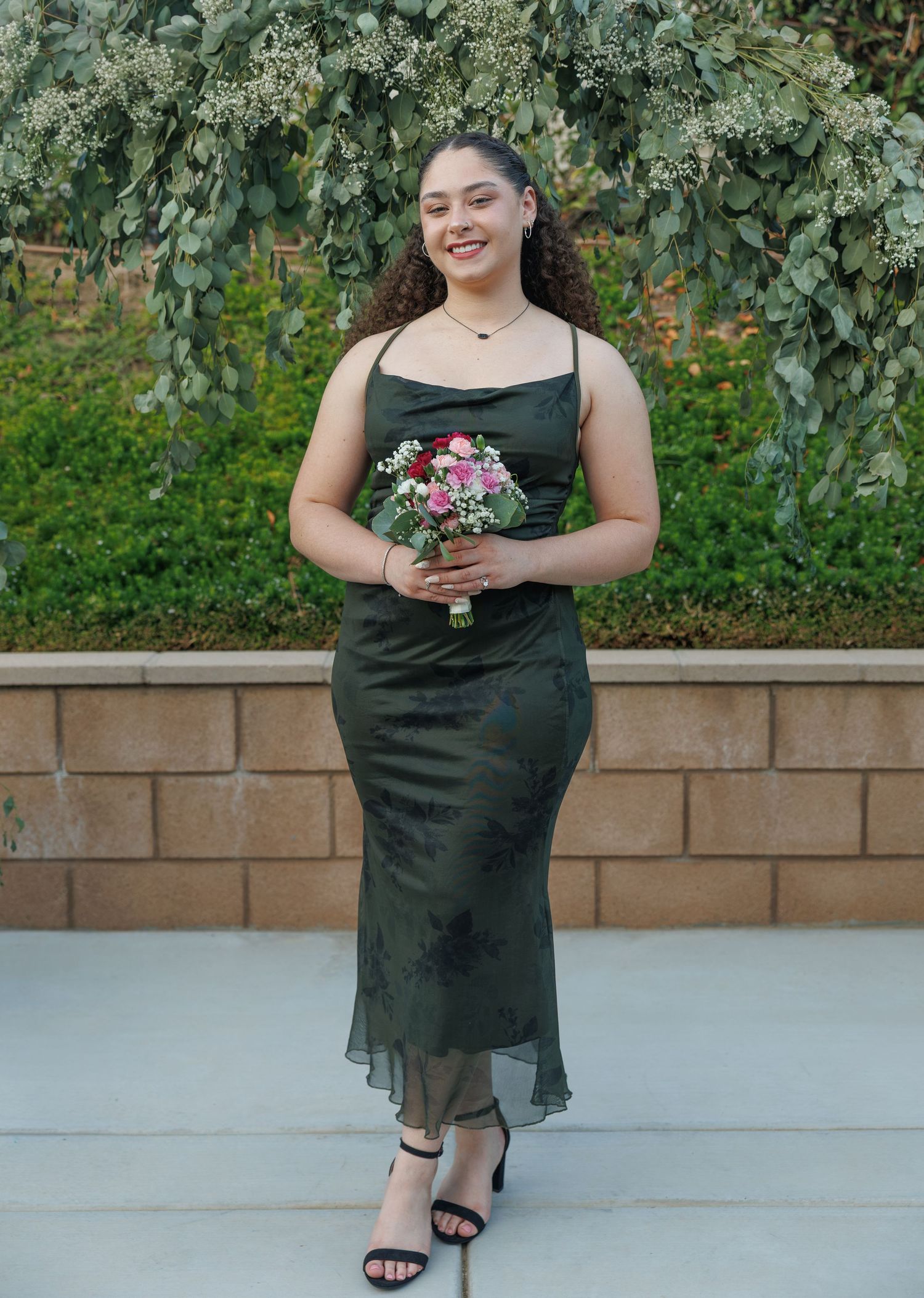 Woman in dark green floral midi dress holding pink bouquet, smiling outdoors near stone wall with greenery.