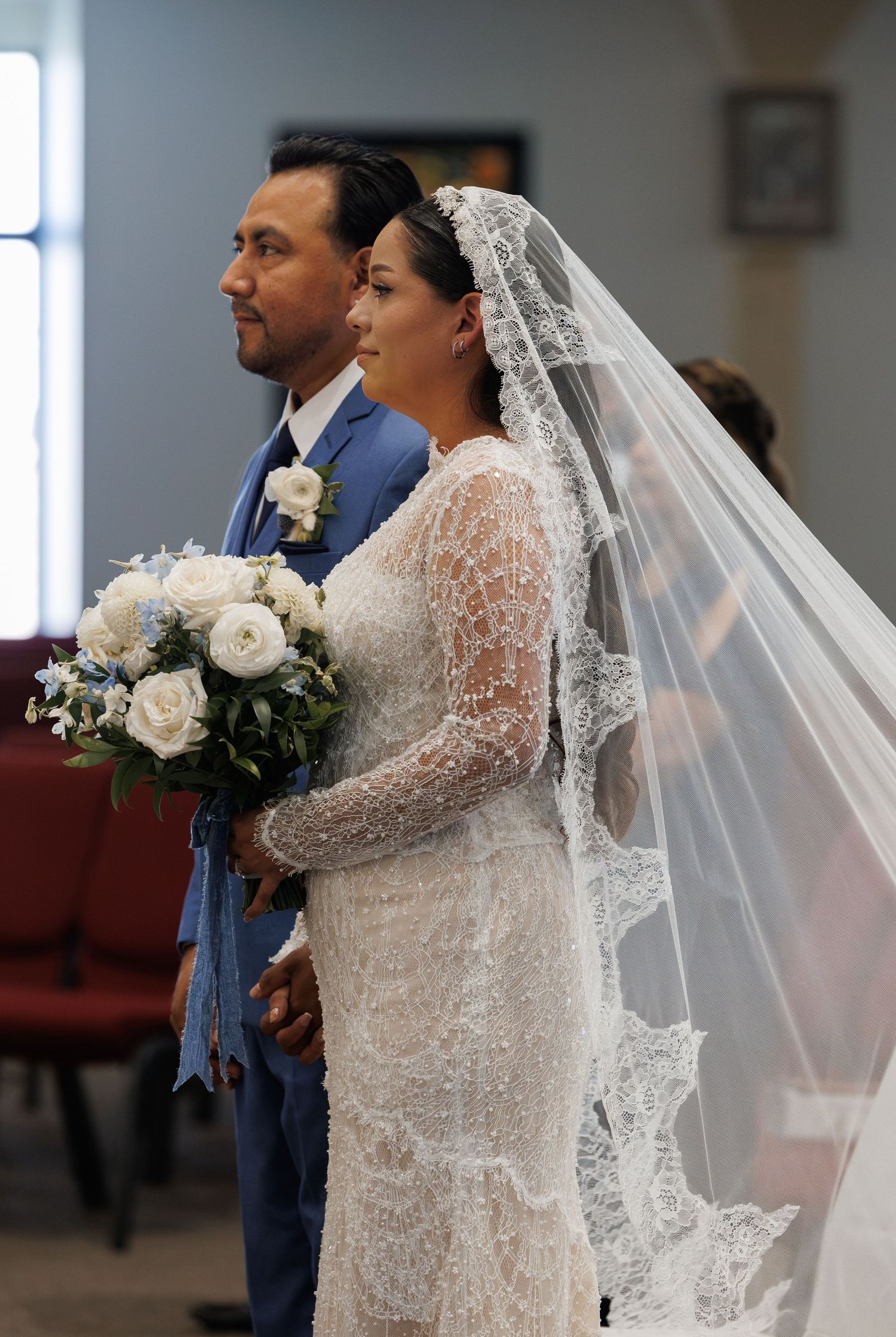 Bride in a beaded lace gown and long cathedral veil holds a white rose bouquet beside her groom in a blue suit during a church ceremony.