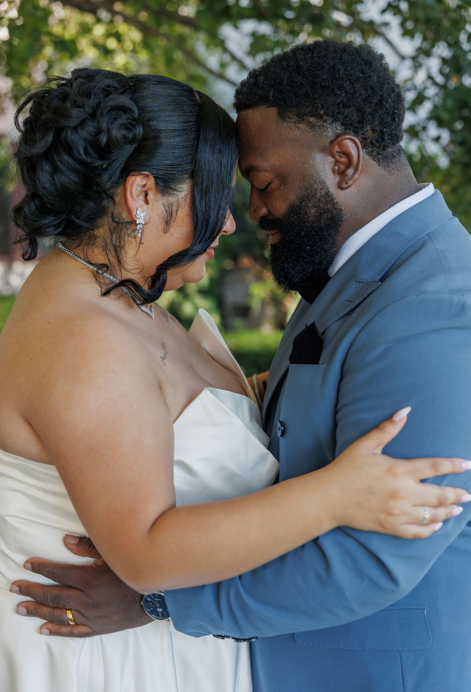 A bride and groom in wedding attire share an intimate forehead-to-forehead moment outdoors, embracing tenderly.
