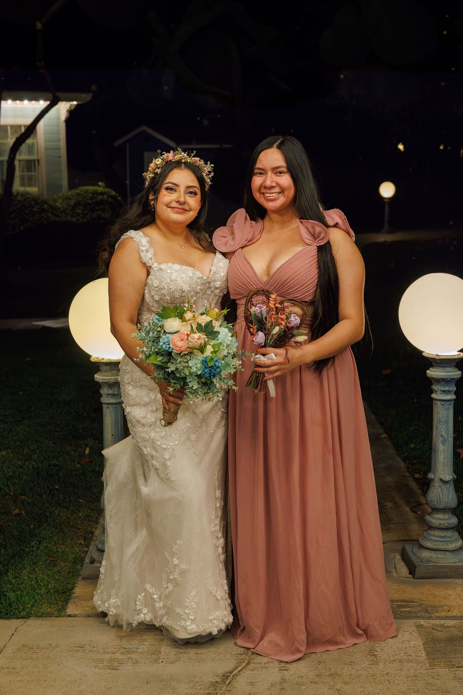 Bride in lace gown with floral crown poses with bridesmaid in dusty rose dress holding bouquets.