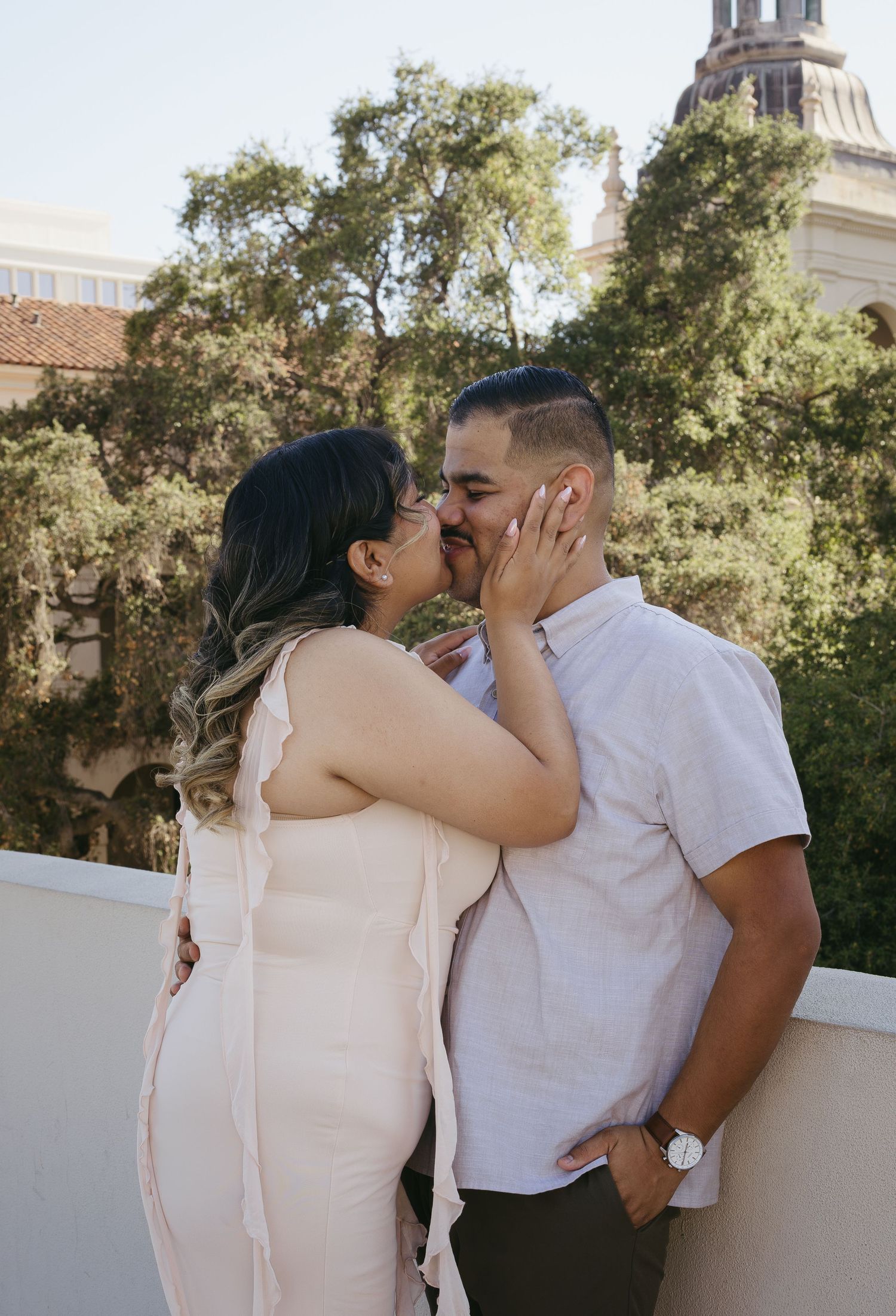 A couple shares a romantic kiss on a balcony surrounded by lush trees and a historic building backdrop.