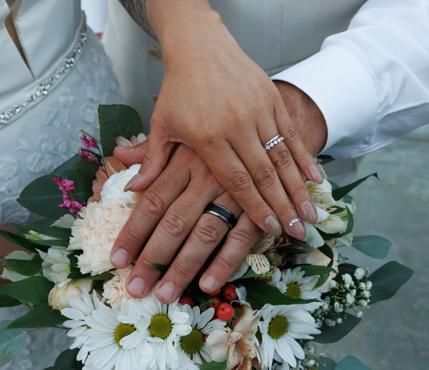 Newlywed couple displaying wedding rings on hands resting over a bridal bouquet of white daisies and roses.