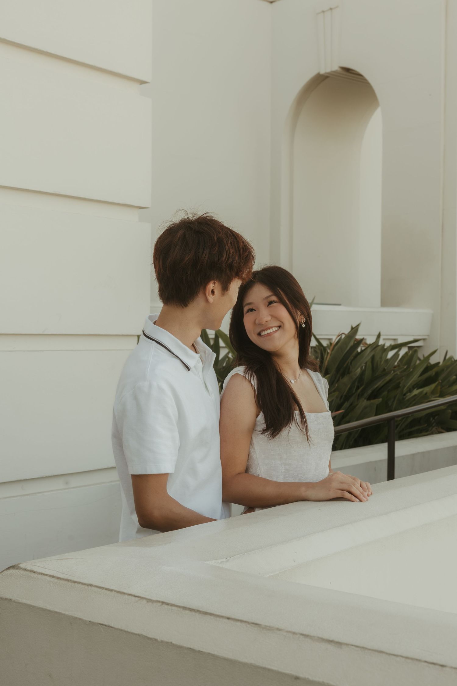 A smiling young Asian couple in white outfits share a tender moment outdoors near a white building.