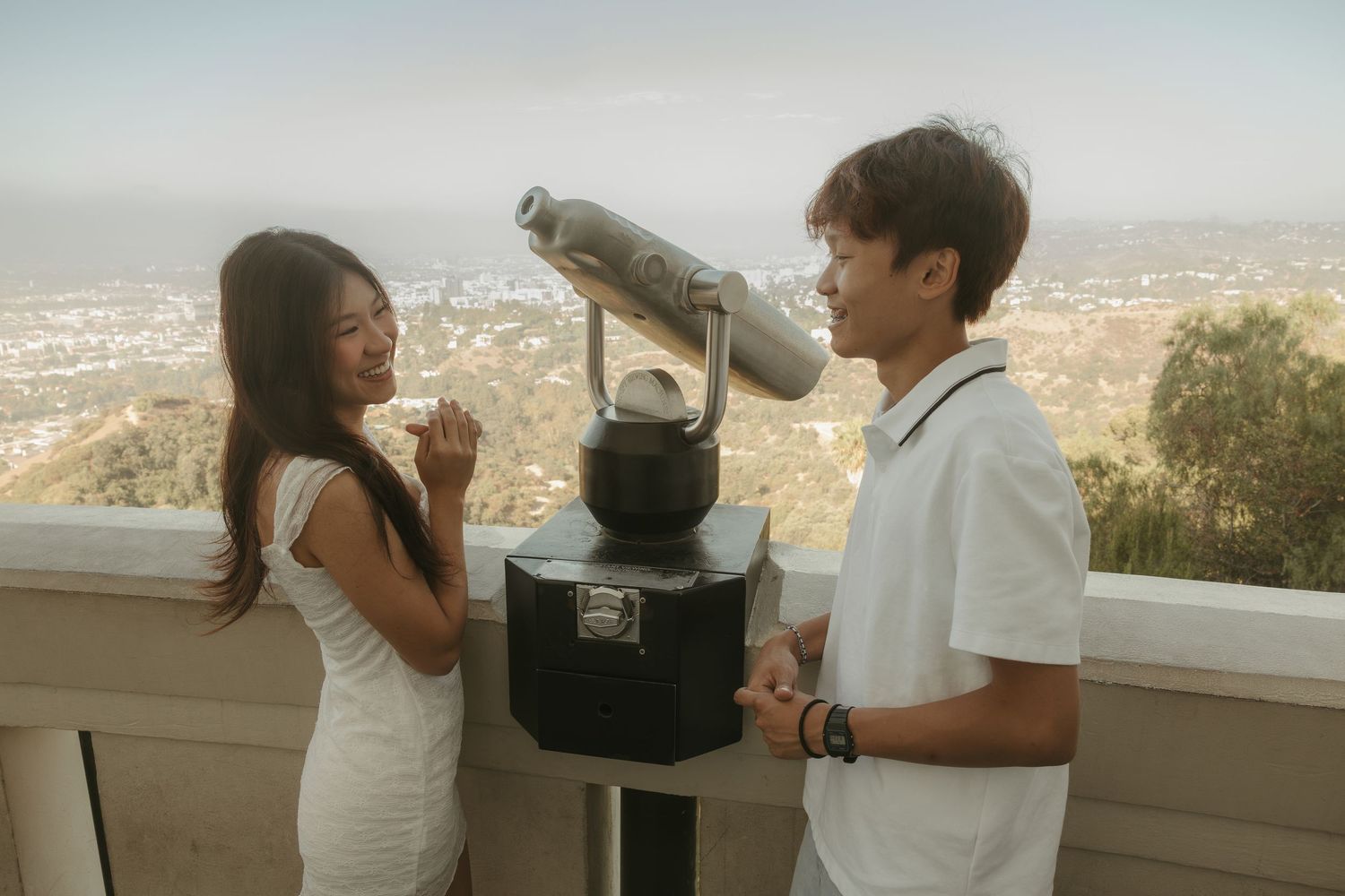A couple stands at a scenic overlook with a coin-operated telescope, enjoying a panoramic city view from a hilltop.