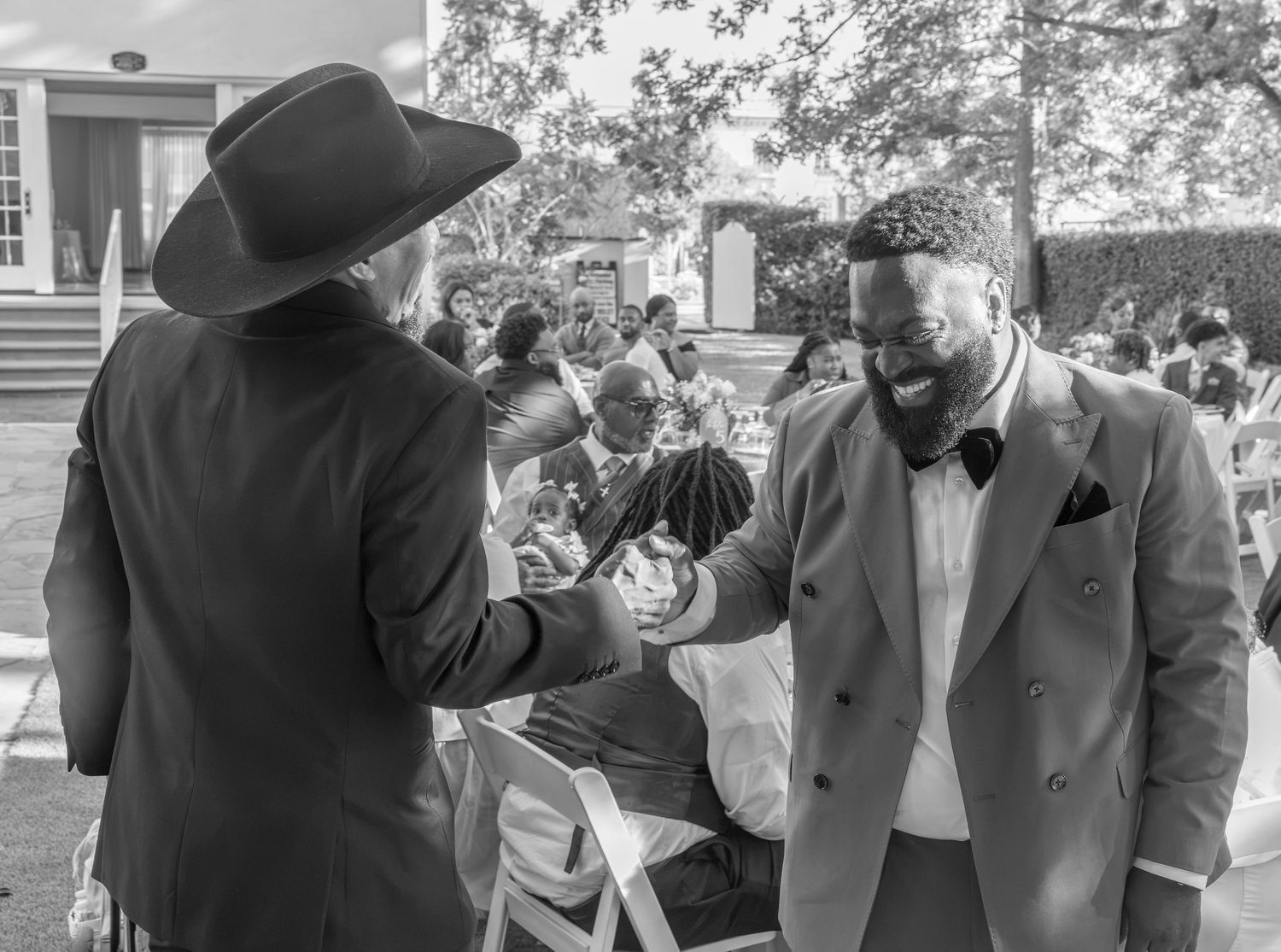 Groom in blue suit greeting guest in black hat at outdoor wedding reception.