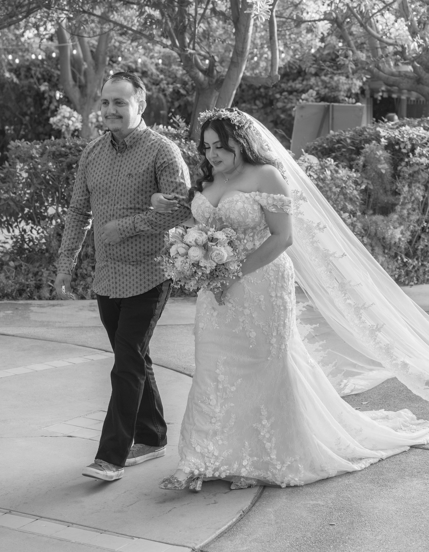 Black and white photo of a bride in lace gown with veil and bouquet walking with a man in patterned shirt.